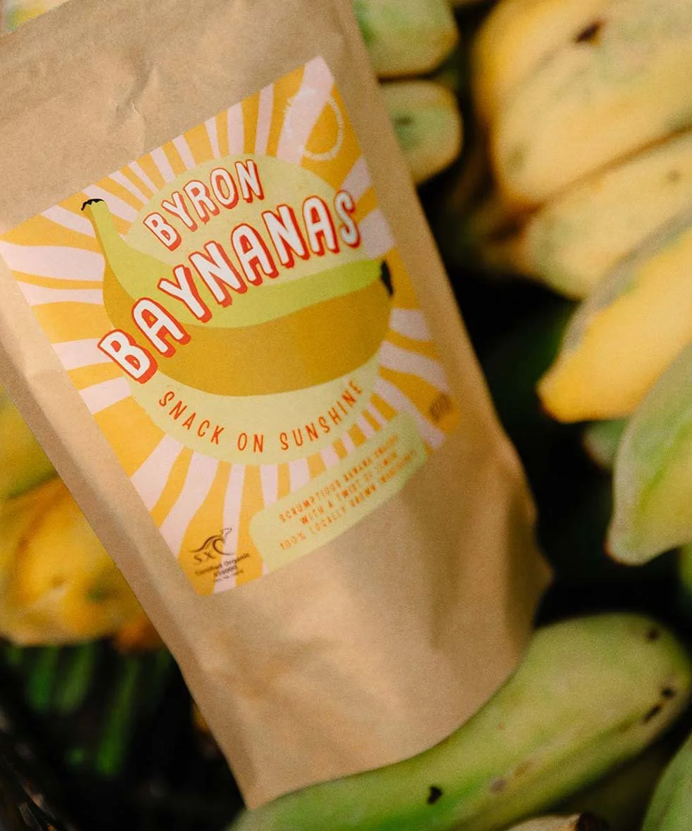 A kraft paper bag labeled 'Byron Bananas' with a sunburst design and a yellow banana illustration, surrounded by ripe yellow bananas.