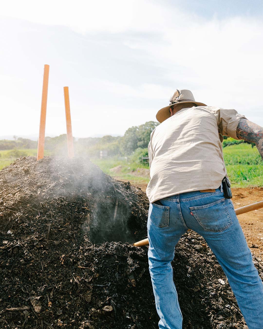 Man wearing a hat and tattoos shoveling burned yard waste in an open field with green crops in the background.