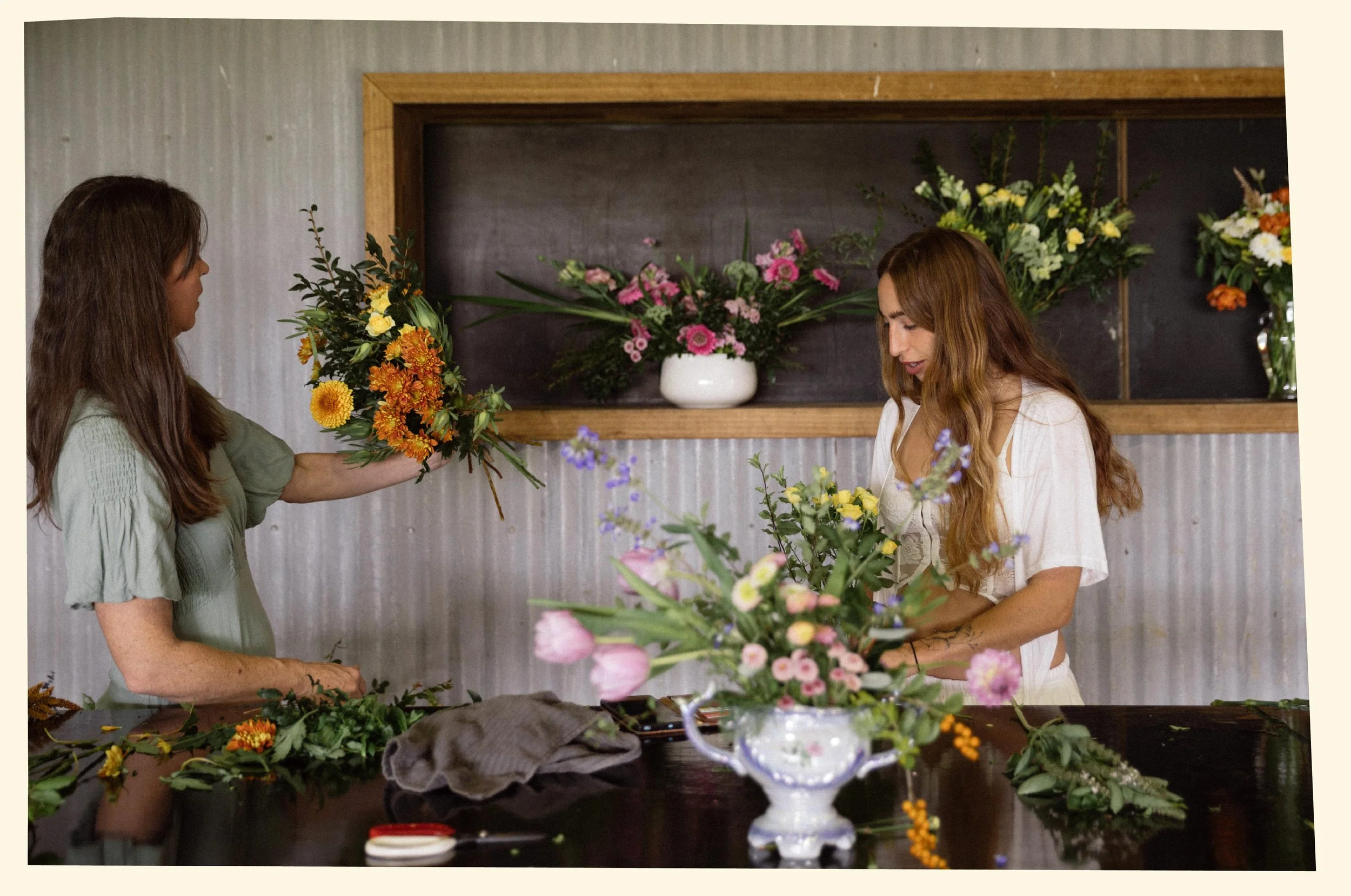 Two women arranging colorful flowers on a table inside a room with a wooden framed wall display of floral arrangements in the background.