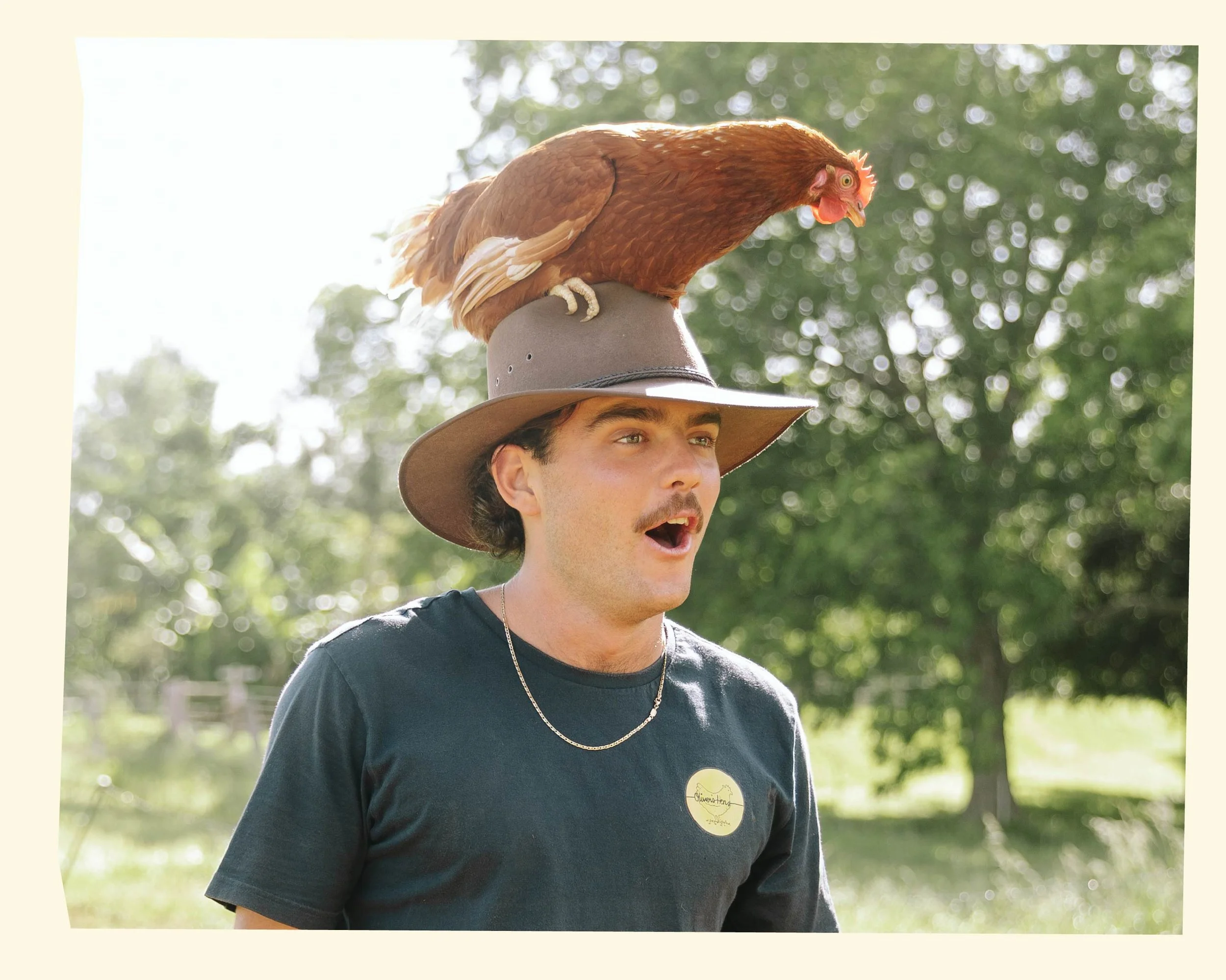 A man wearing a wide-brimmed hat and a black t-shirt with a round gold sticker, standing outdoors with green trees in the background, a chicken perched on his hat.