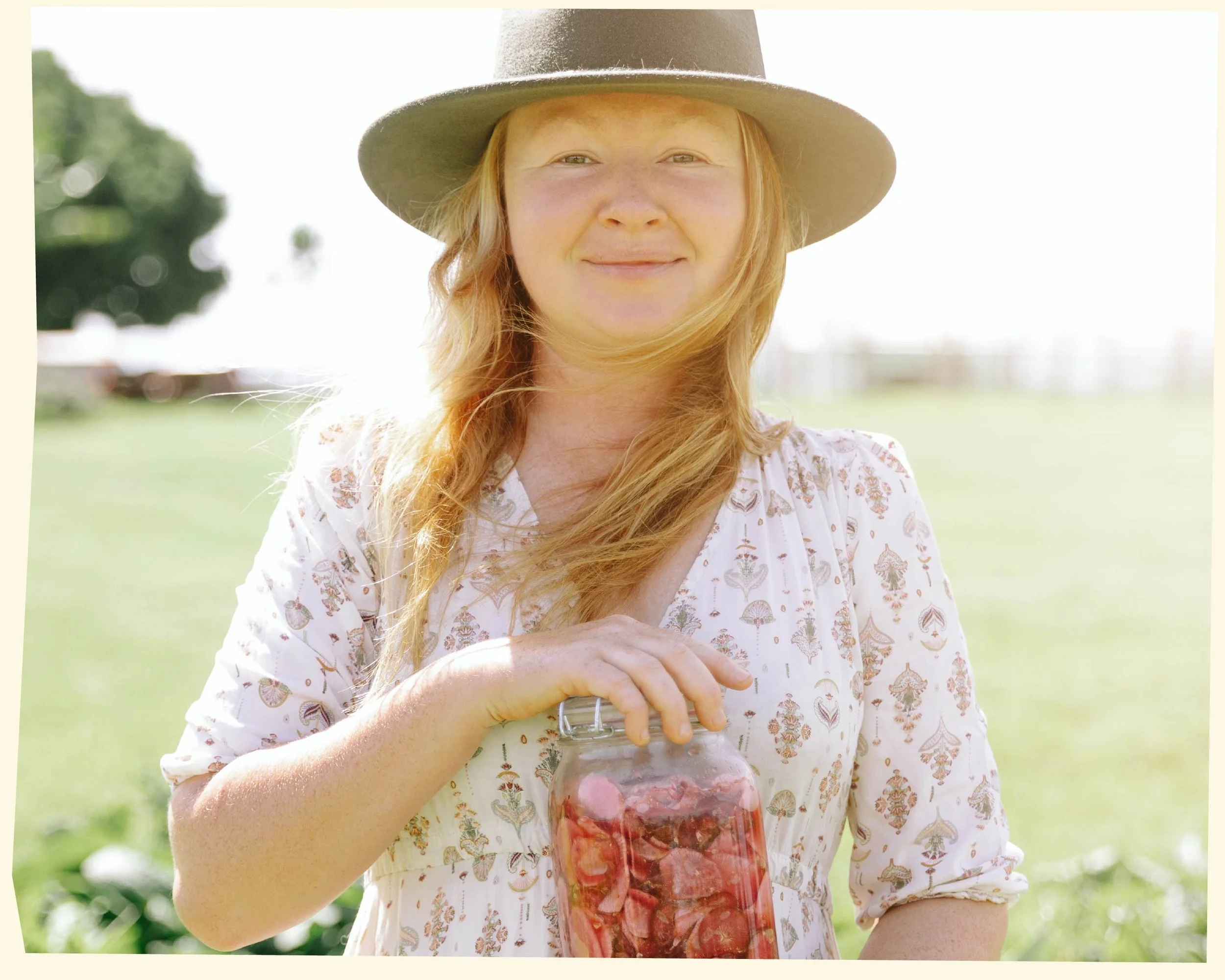 A woman with red hair wearing a wide-brimmed gray hat and a white dress with a floral pattern, outdoors in a green field, holding a jar of fruit or berry preserve.