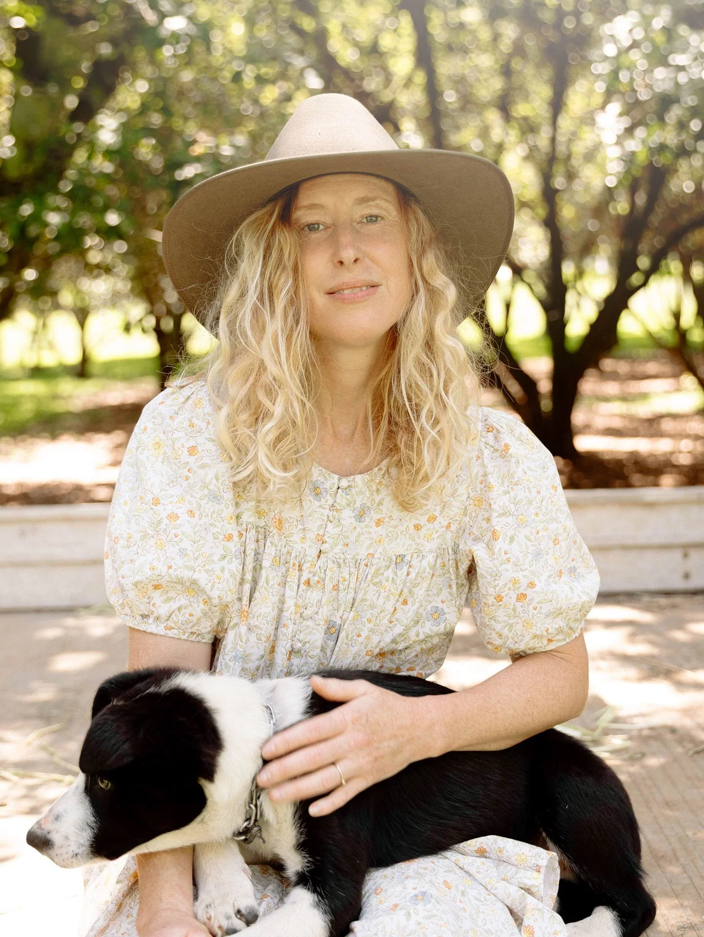 A woman with long blonde hair wearing a wide-brimmed tan hat and a yellow floral dress sitting outdoors with a black and white puppy in her lap.