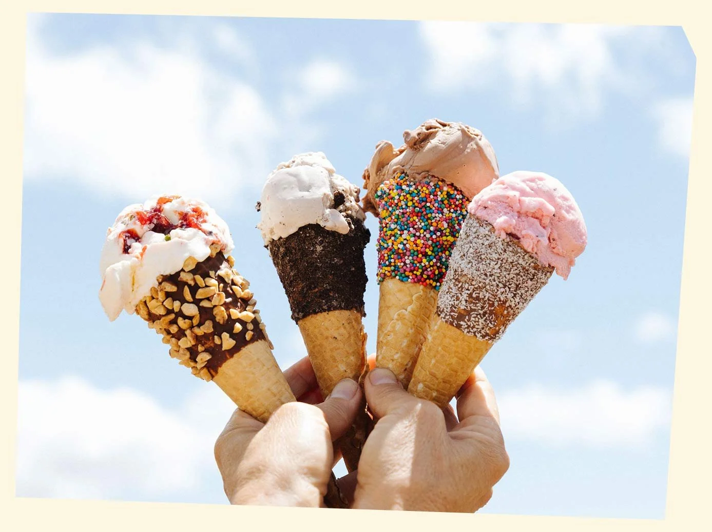 Four hands holding different ice cream cones with a blue sky and clouds background.