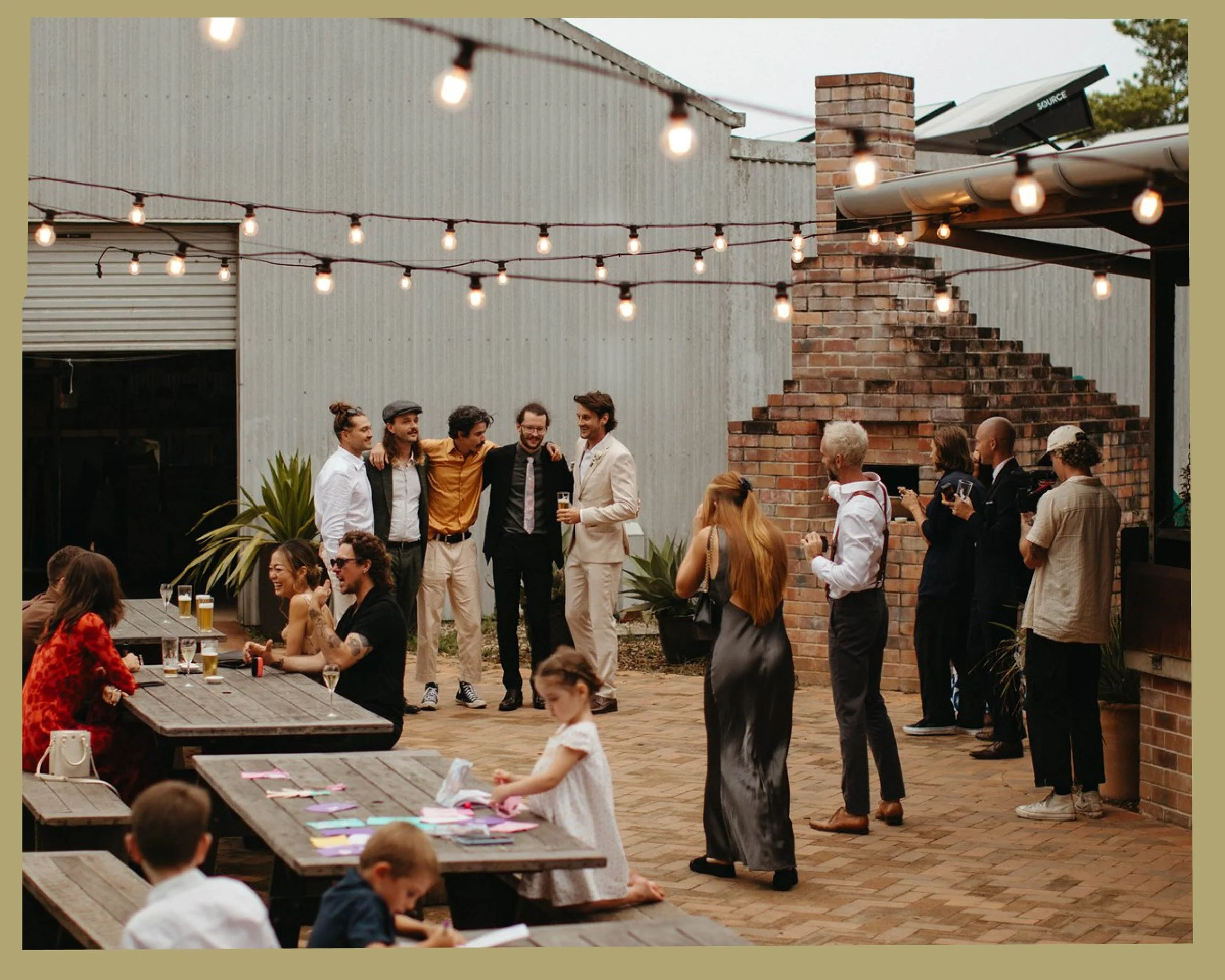 A diverse group of people socializing at an outdoor event under string lights, with a brick chimney, corrugated metal wall, and solar panels in the background.