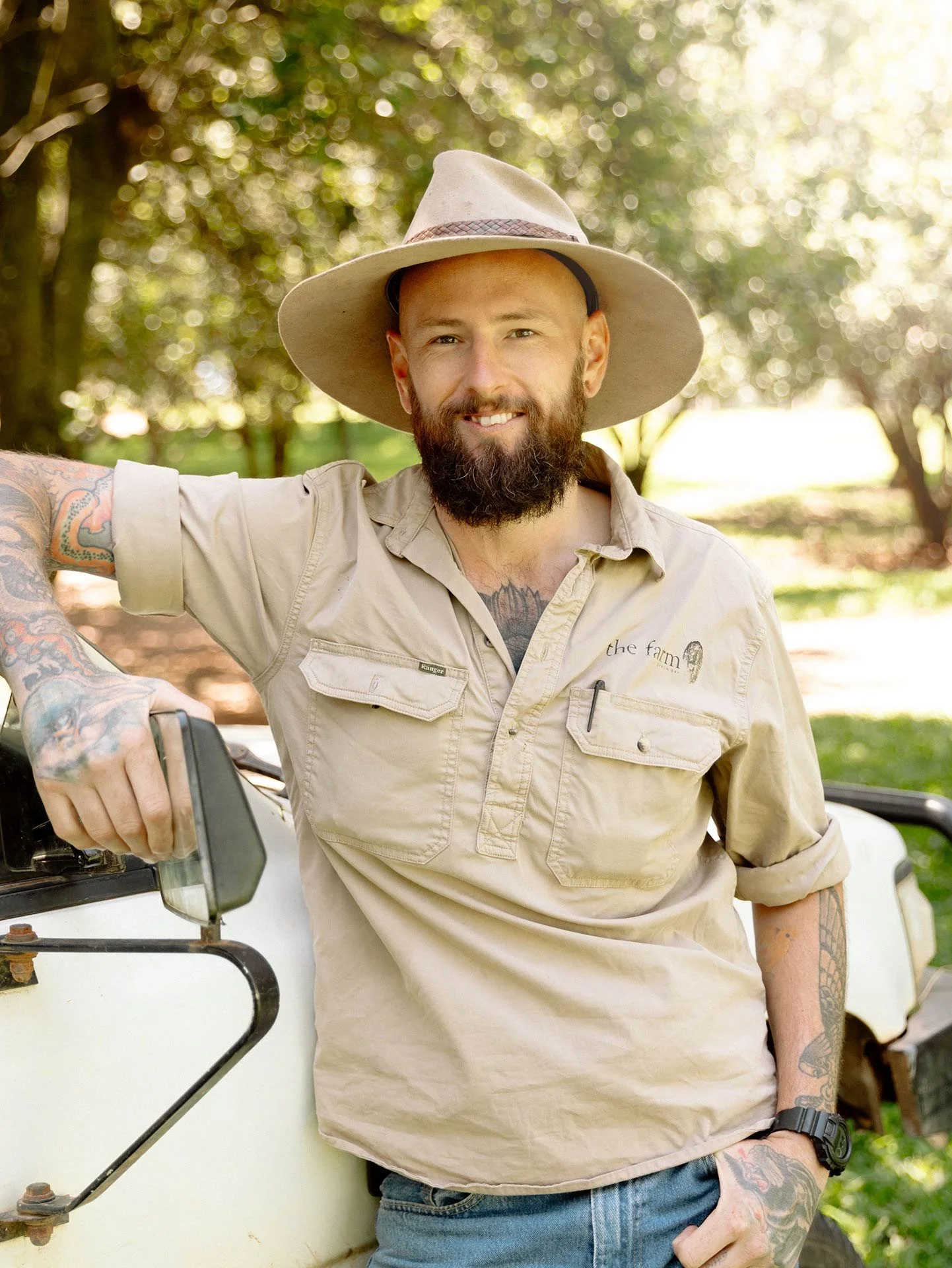 A bearded man with tattoos wearing a wide-brimmed hat and khaki shirt, smiling and standing outdoors next to a white utility vehicle.