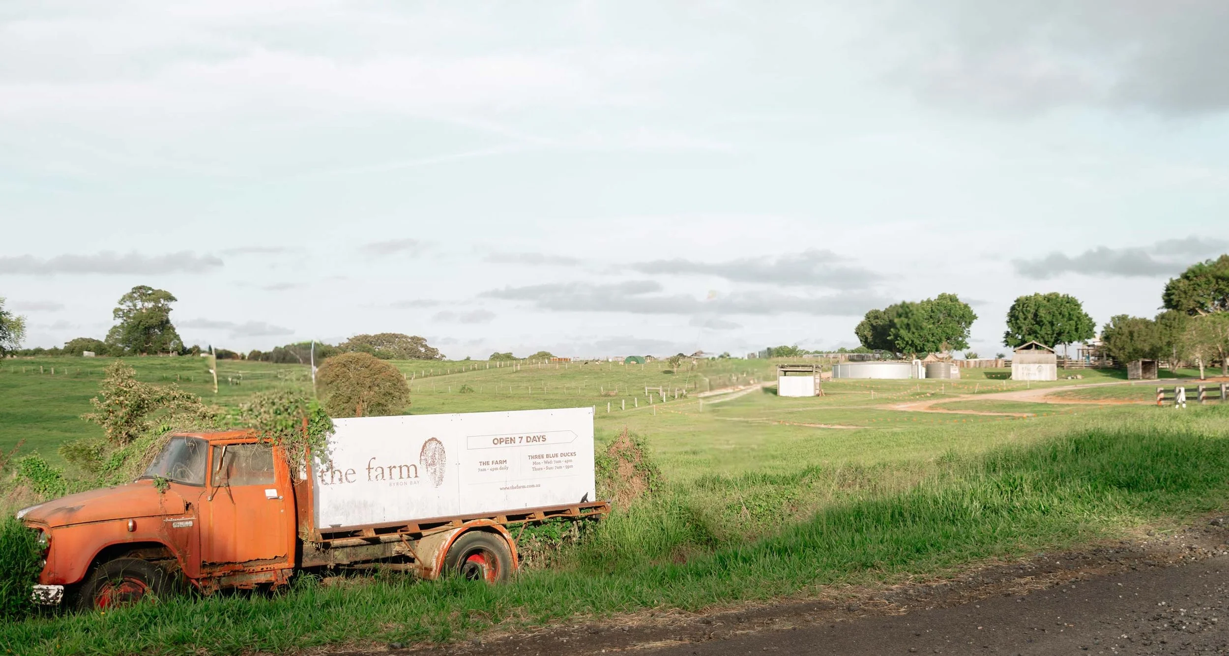 A rustic orange truck parked on a grassy field with a white sign on the side advertising The Farm Byron Bay, open seven days a week. The background features a vast green field with scattered trees, small buildings, water tanks, and cloudy sky.