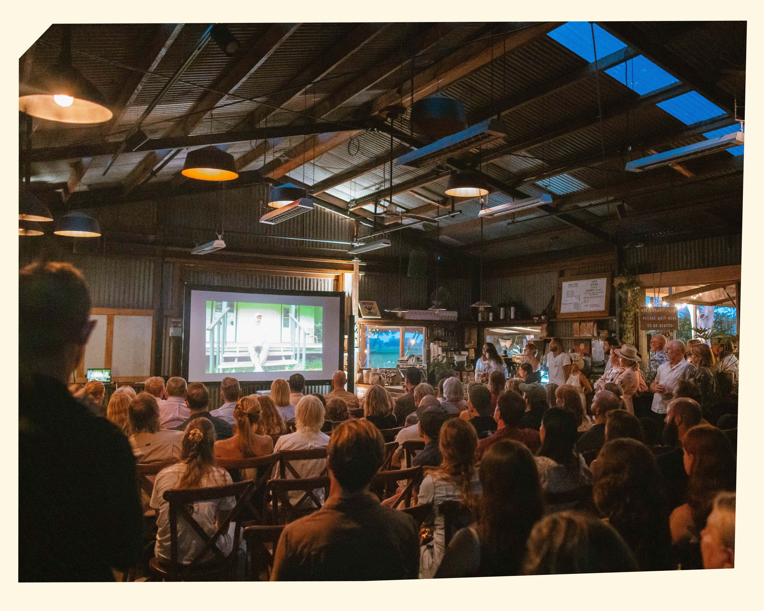An indoor gathering with many people seated and standing, watching a presentation or performance on a large screen, in a room with a high wooden ceiling with skylights and hanging lights.