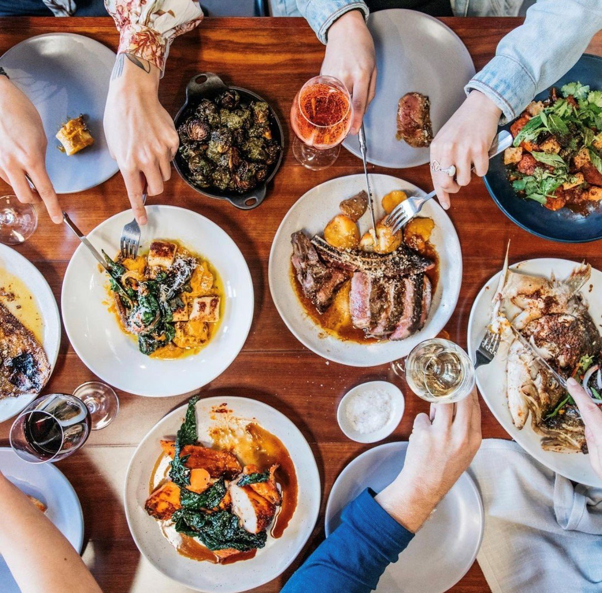 Overhead view of a group of people enjoying a meal with various dishes including steak, pasta, vegetables, and drinks on a wooden table.