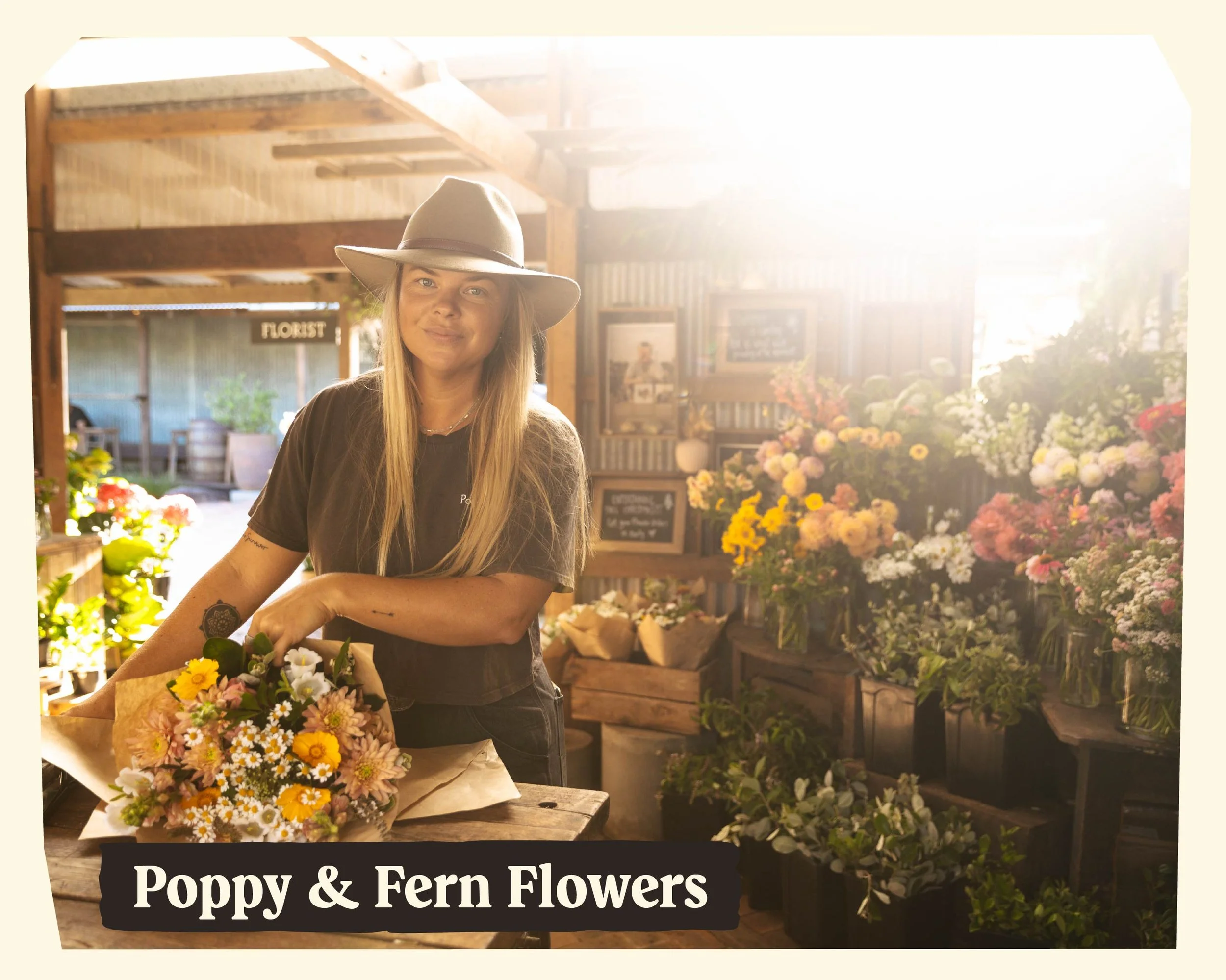 A woman with long blonde hair wearing a beige wide-brimmed hat and a dark t-shirt standing in front of a flower stand filled with colorful potted flowers, with a sign reading "Poppy & Fern Flowers" in the foreground, sunlight illuminating the scene.