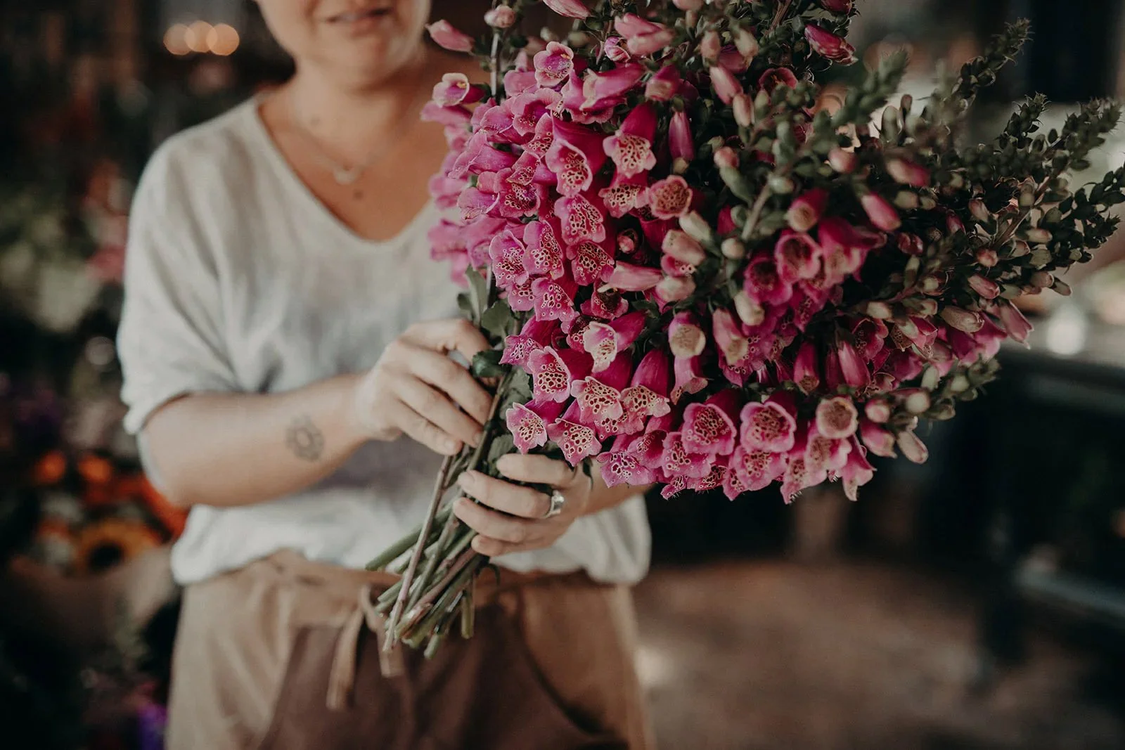 A person holding a large bouquet of pink and purple flowers, with a blurred background of a flower shop or florist's workspace.