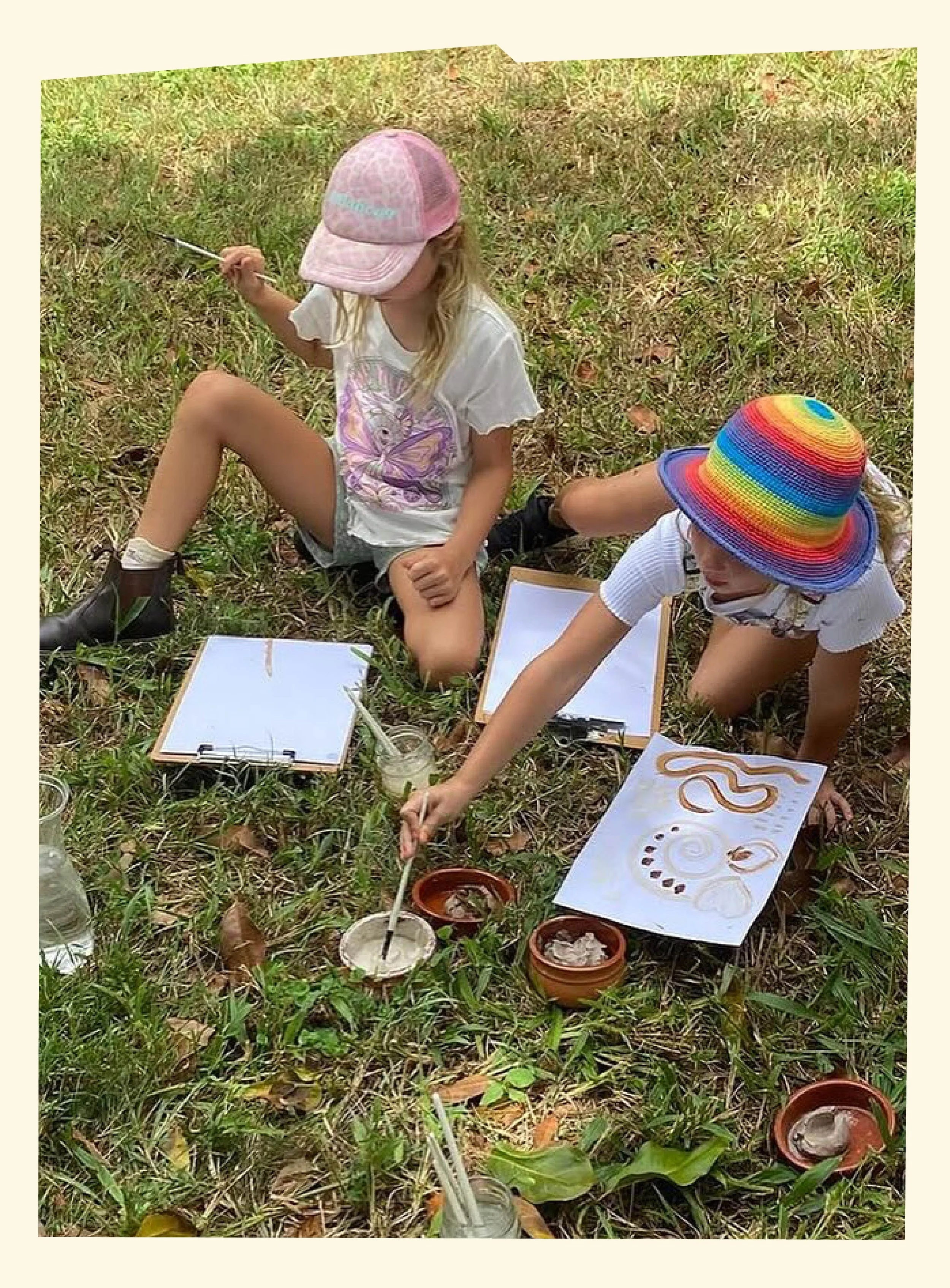 Two children sitting on grass outdoors, painting and drawing with art supplies, including jars of paint, paper, and brushes. One child wears a pink cap, and the other wears a rainbow-colored sunhat.