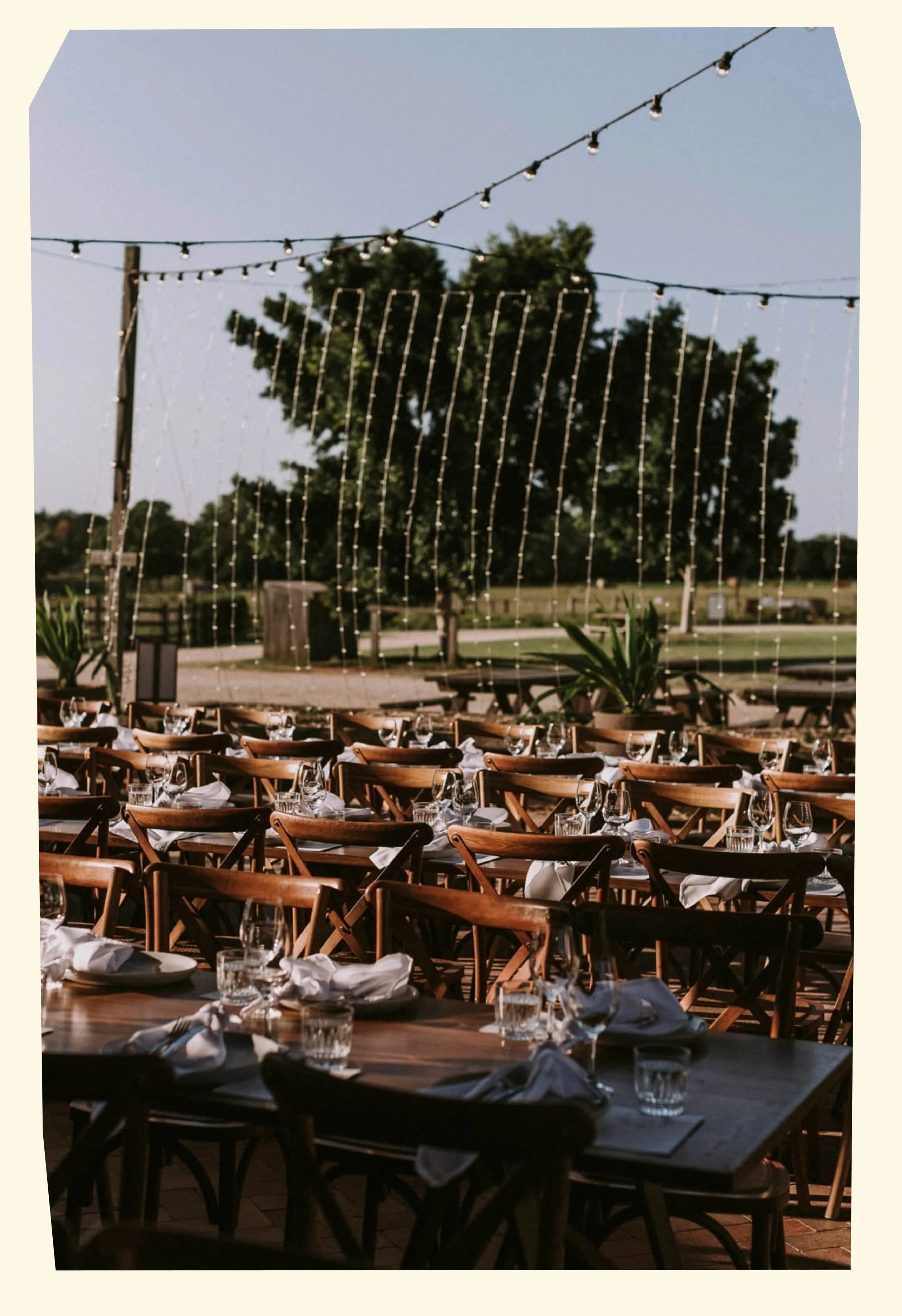 Outdoor restaurant setup with wooden tables and chairs, set with glasses and napkins, under string lights, with trees and a clear sky in the background.
