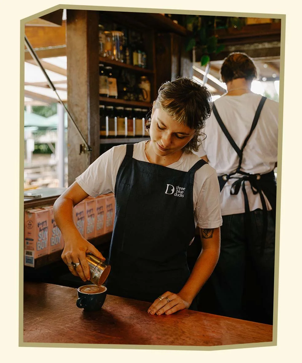 A woman pouring steamed milk into a coffee cup, creating latte art, while wearing a black apron with the logo 'three blue ducks', in a cozy café setting with a barista in the background.