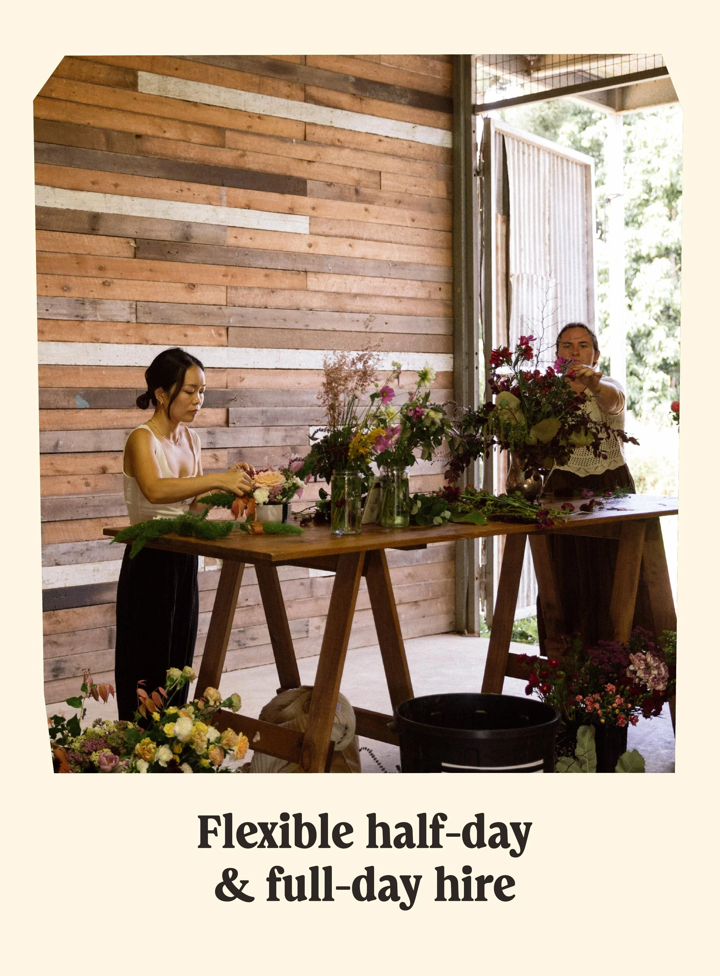 Two women arranging flowers on a wooden table inside a rustic building with a wooden wall and open door leading outside.