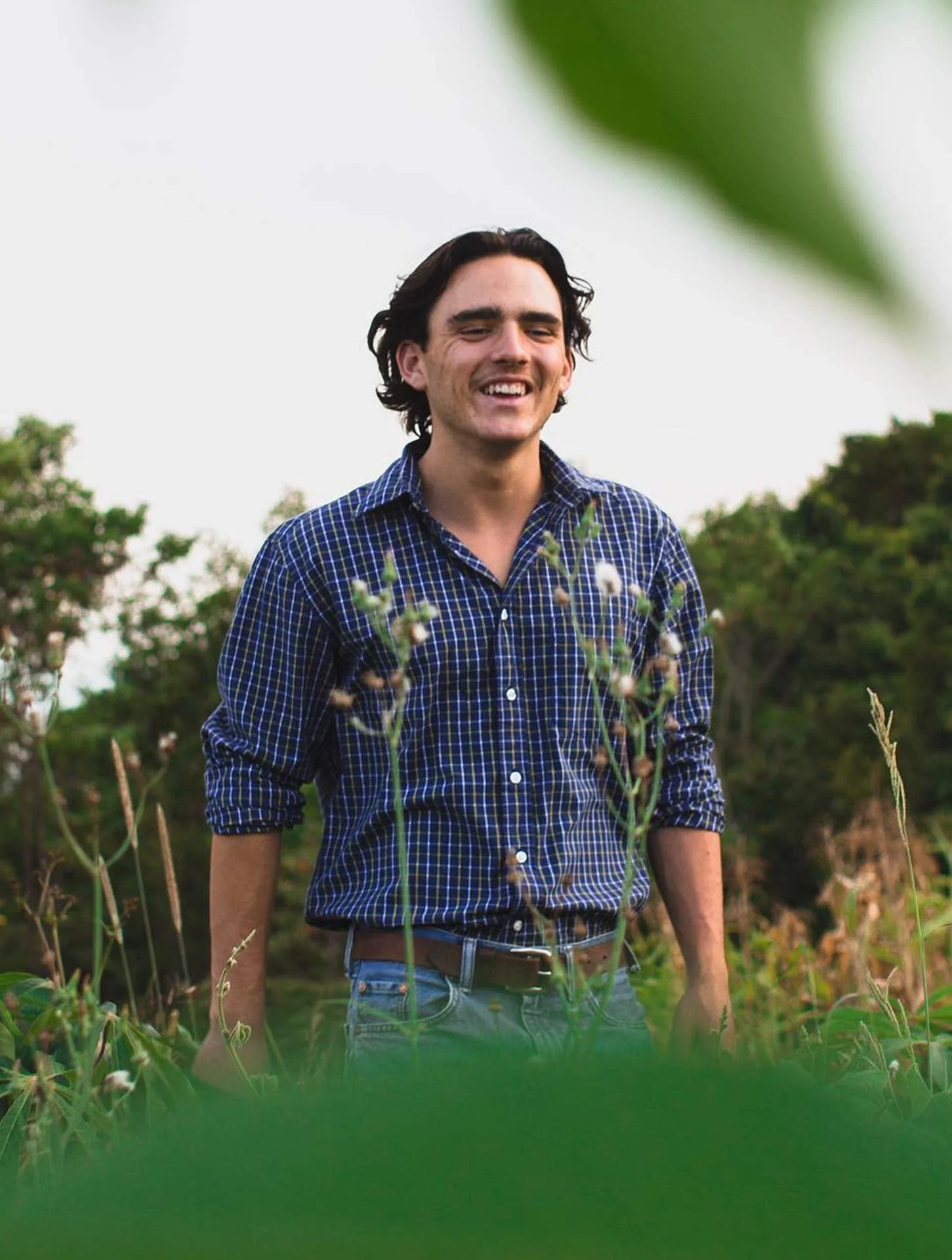 A young man with dark wavy hair, wearing a blue checkered shirt with the sleeves rolled up, stands outdoors in a field with plants and trees in the background. He is smiling and appears to be enjoying himself.