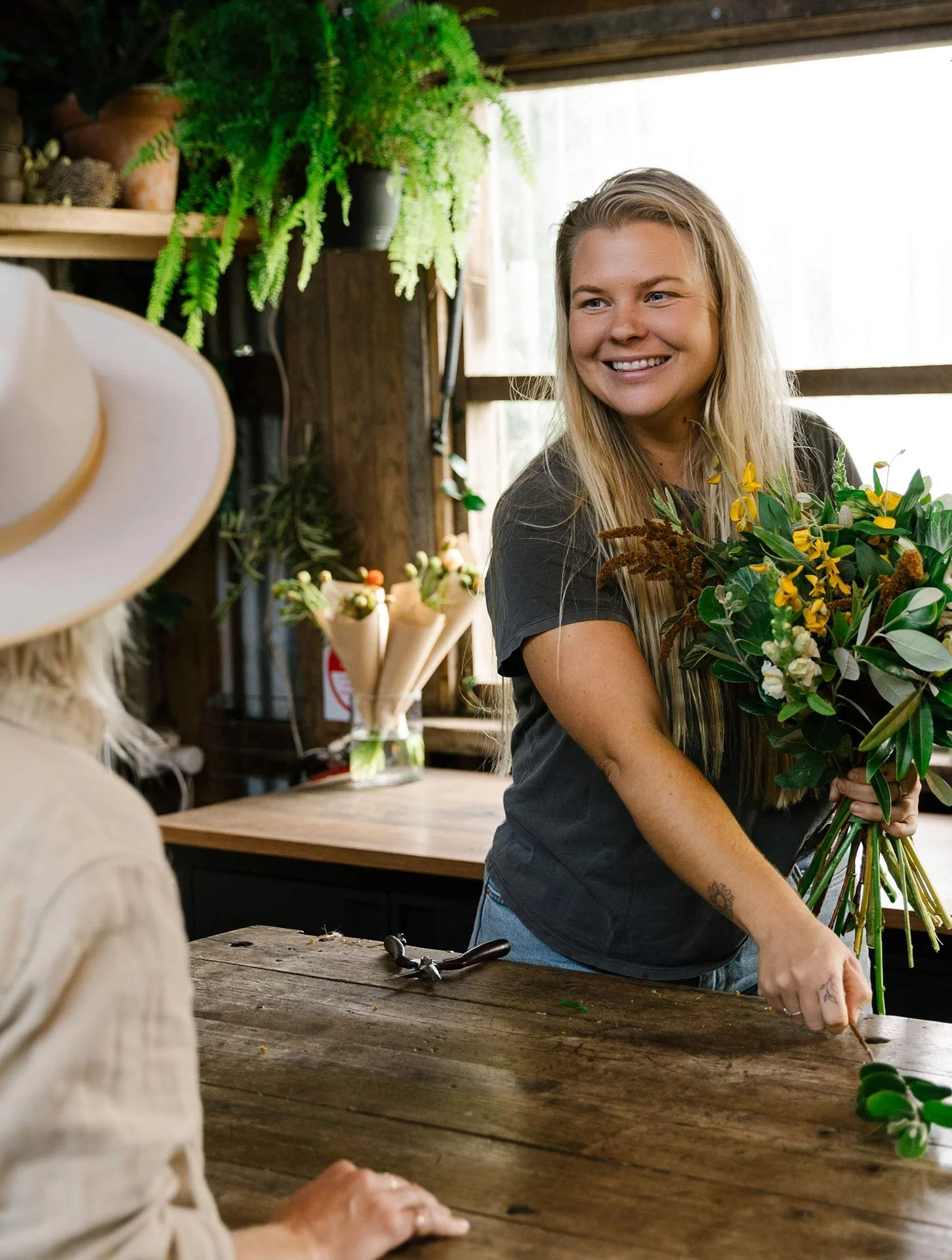 A woman in a dark t-shirt holding a bouquet of yellow and green flowers, smiling indoors with plants and wood decor, a hat on the left, and gardening tools on the table.
