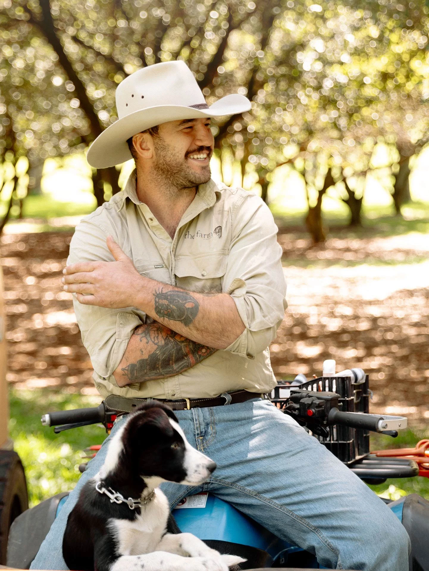 A smiling man wearing a cowboy hat and beige shirt, sitting outdoors with a black and white puppy resting on his lap. The background shows trees and sunlight.