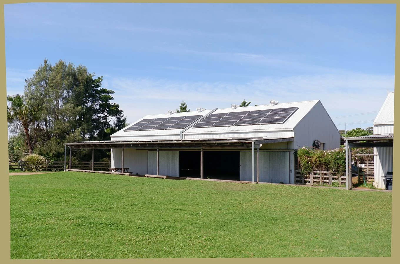 A white barn with solar panels on the roof, surrounded by green grass and trees under a blue sky.