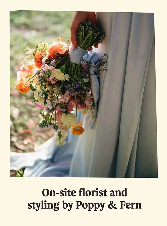A person holding a cascading bouquet of mixed flowers, including orange, pink, purple, white, and green blooms, in front of a draped curtain at an outdoor event with a grassy background.