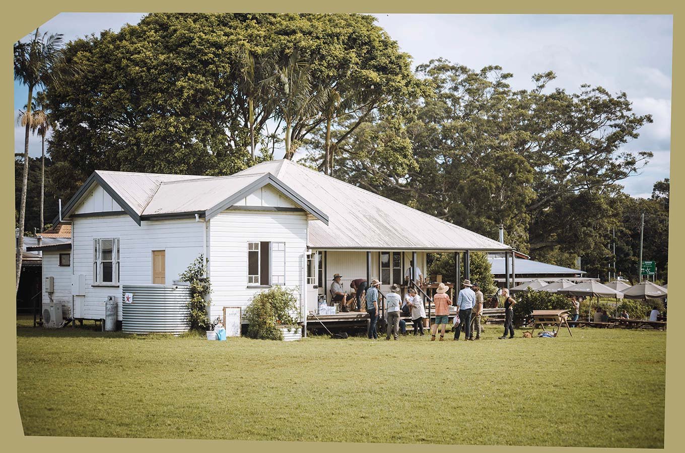 A group of people gathered on the porch of a white wooden house with a metal roof, surrounded by large trees and a green lawn, in a rural setting.