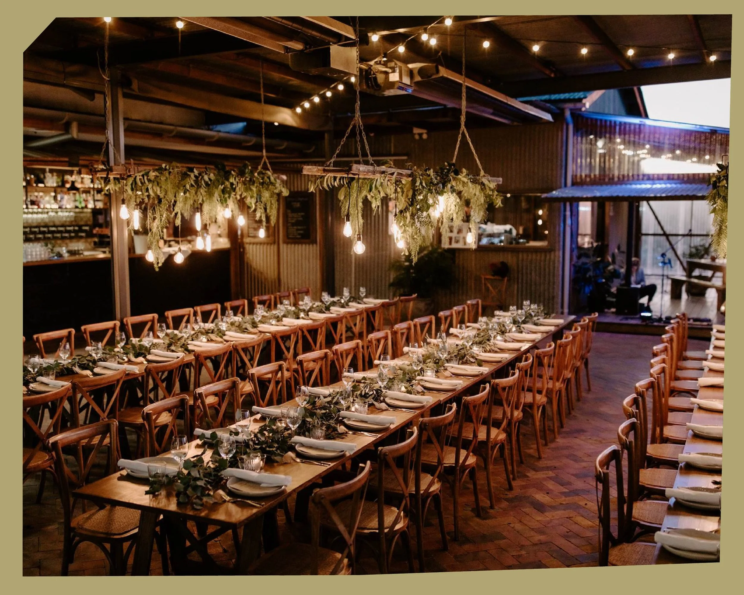 Indoor celebration venue with long wooden tables set with white plates, glasses, and greenery centerpieces, decorated with hanging lights and hanging greenery, and a bar area in the background.