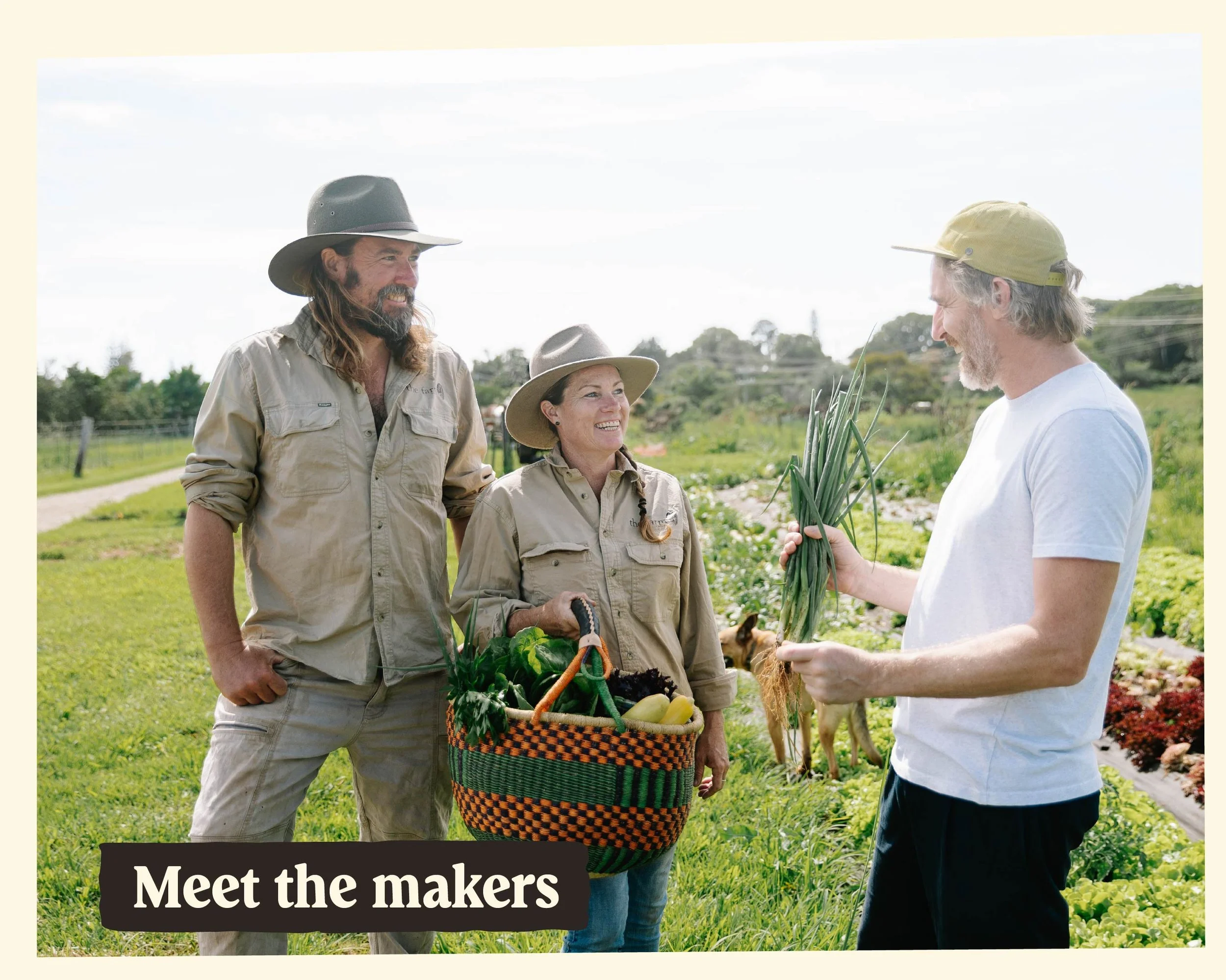Three people, two men and one woman, are outdoors in a garden or farm setting. They are dressed in casual outdoor clothing and hats, and are engaged in conversation. The man on the right is holding a bundle of green onions, and the woman in the middle is holding a basket filled with vegetables, including a zucchini and carrots.