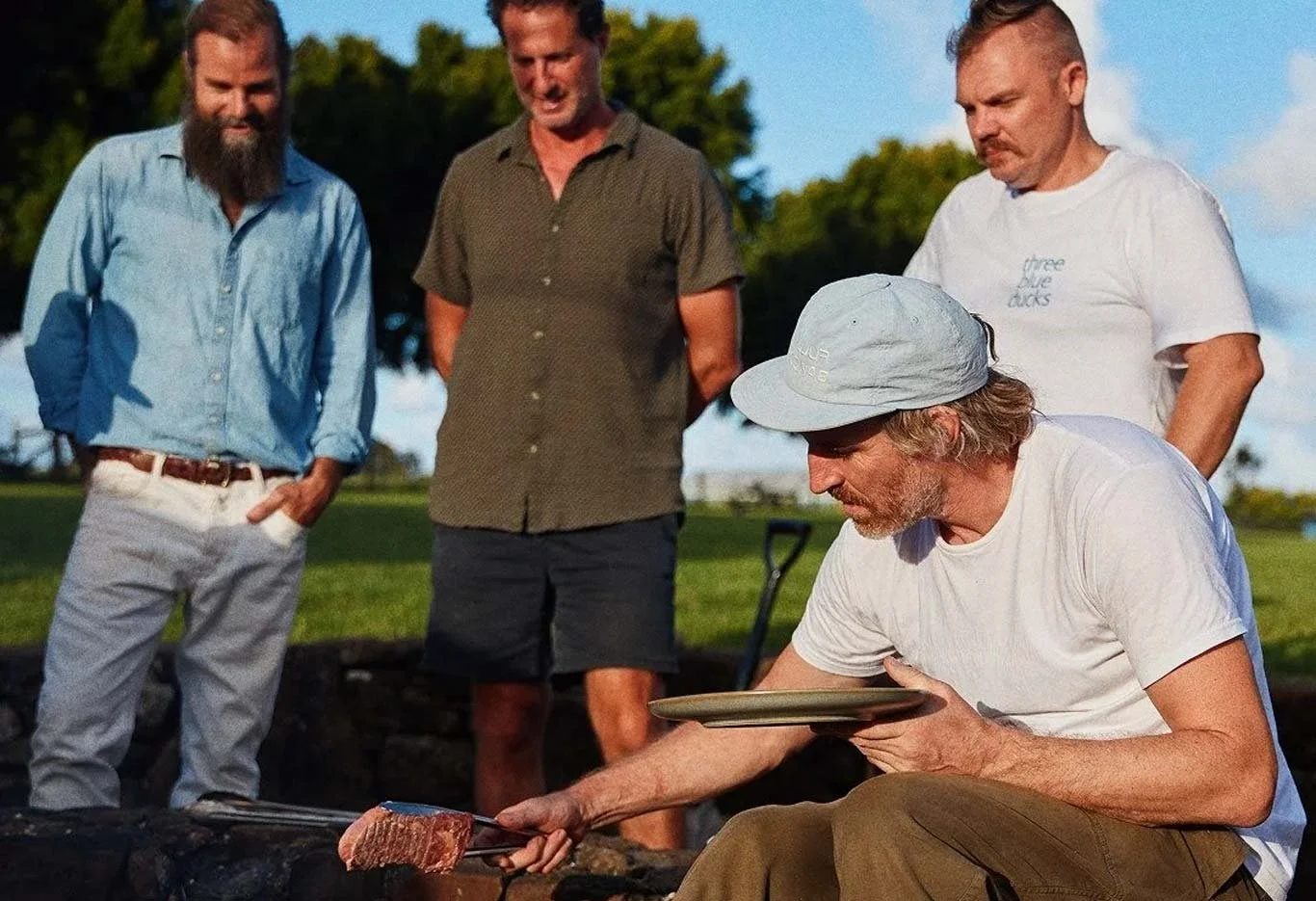 Four men outdoors, one sitting and handling a piece of meat with tongs, others watching, during a barbecue in the evening with a clear sky and trees in the background.