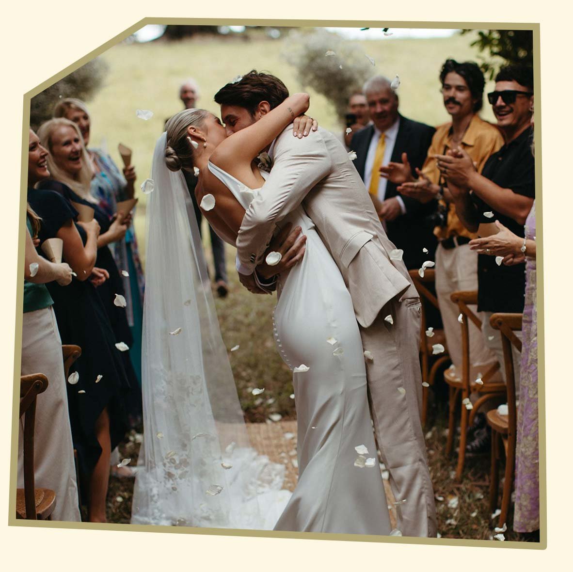A happy bride and groom kiss while guests celebrate at an outdoor wedding with flower petals falling.