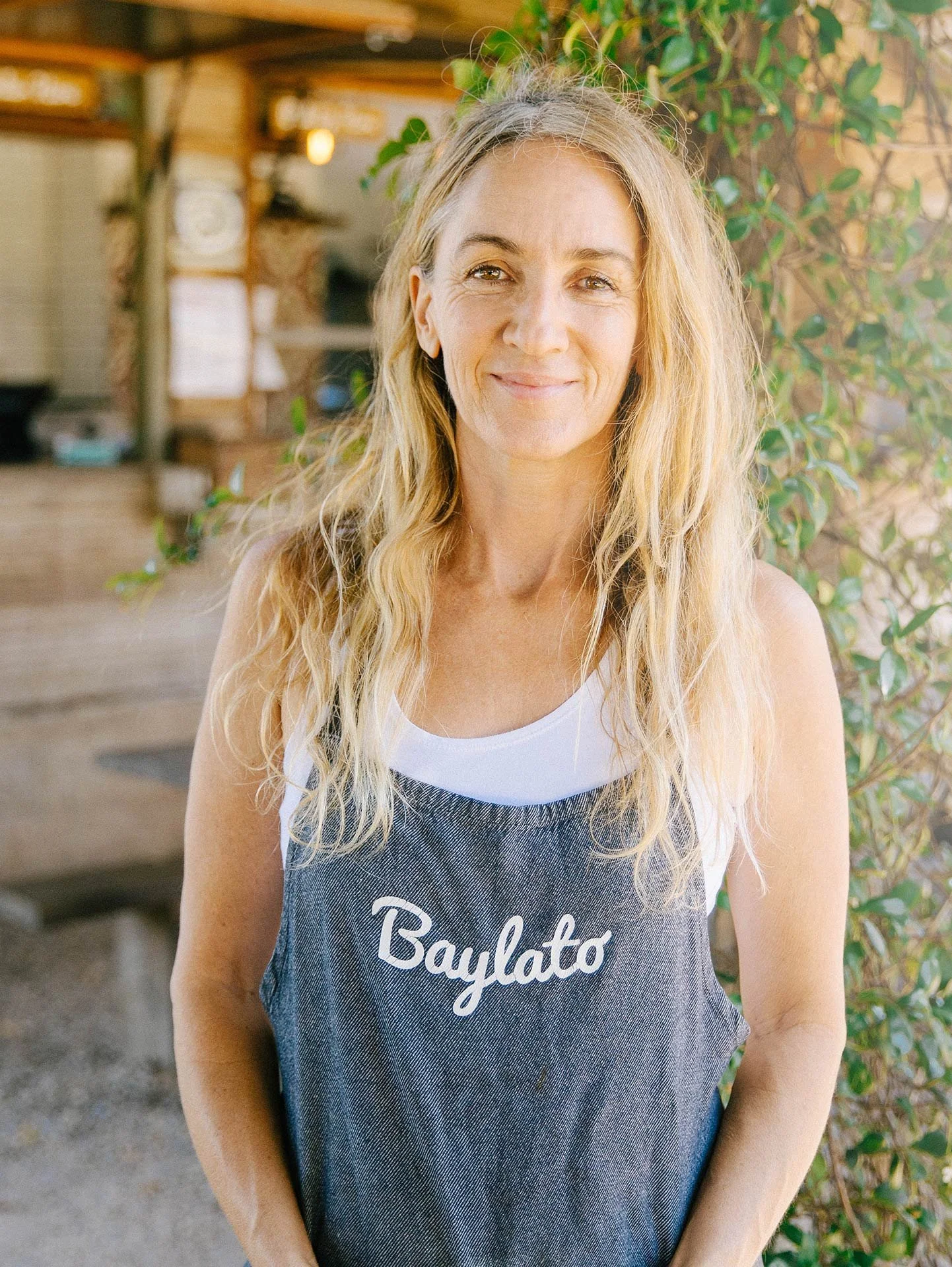 A woman with long blonde curly hair is smiling and standing outdoors near green bushes, wearing a sleeveless shirt that says 'Baylato'.