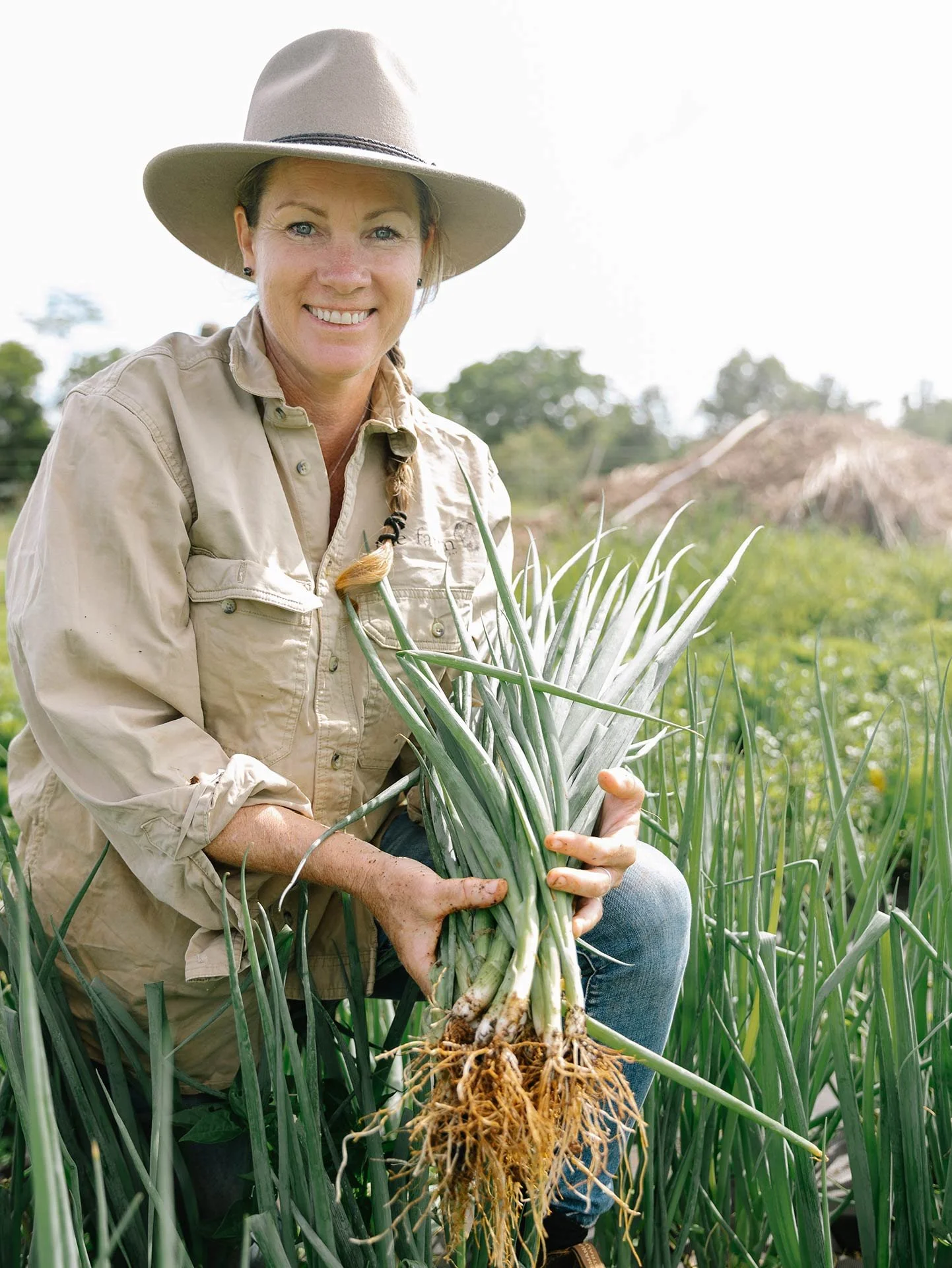 A woman in outdoor work clothes and a wide-brimmed hat kneels in a field of green onions, holding freshly harvested onions with roots attached, smiling at the camera.