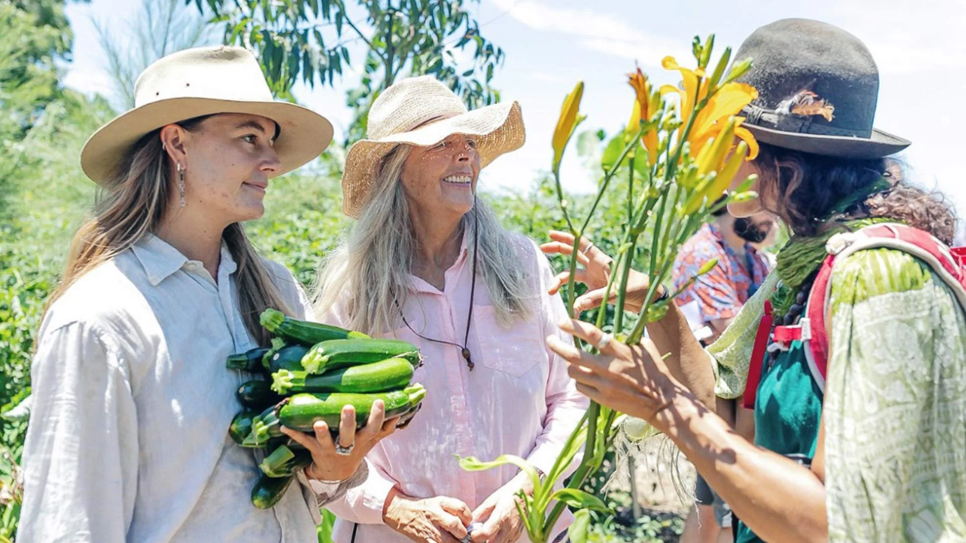 Three women in hats exchanging flowers and vegetables during a sunny outdoor event in a lush green field.