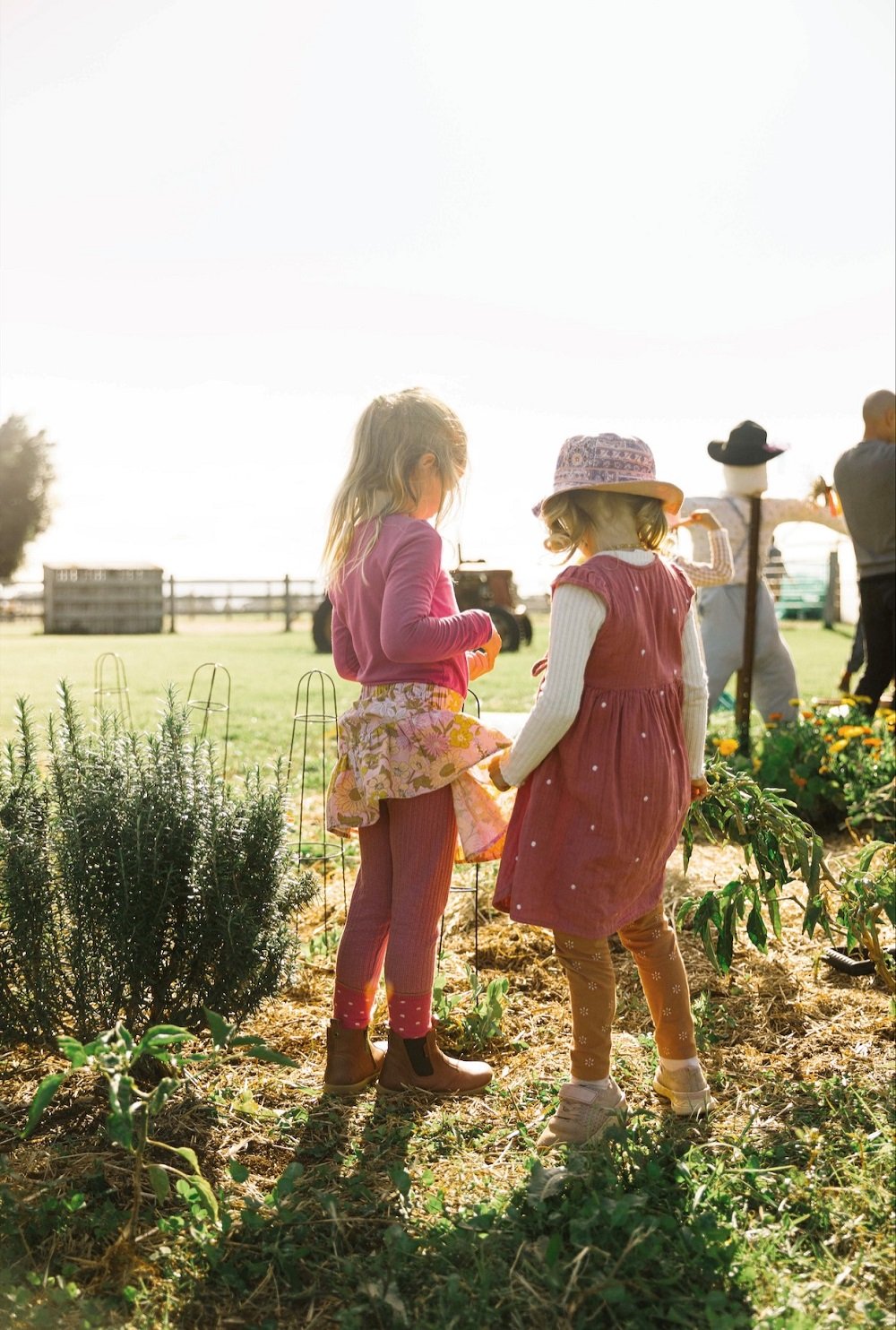 Two young girls in colorful clothing and hats, standing in a garden or farm, engaging in gardening or harvesting, with sunlight and other people in the background.