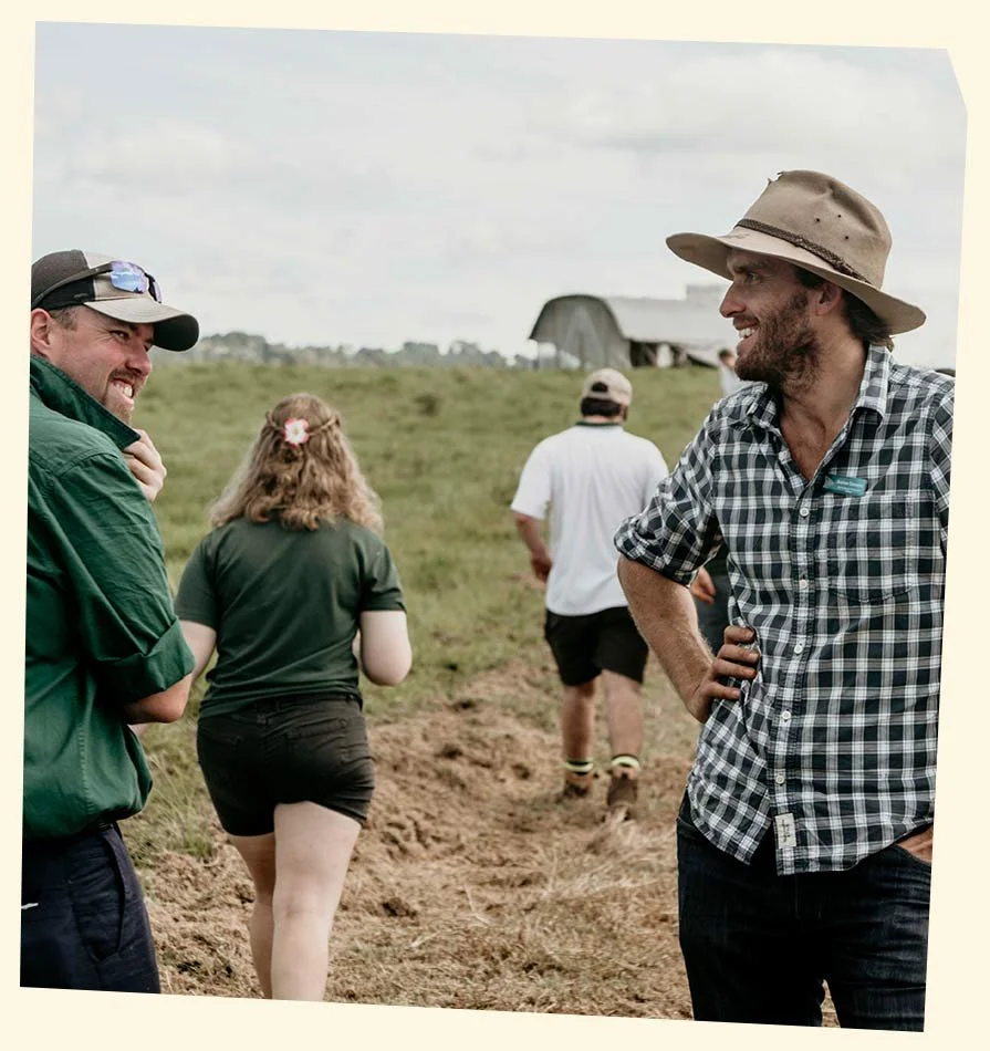 People walking on a farm or countryside, with two men in the foreground smiling and talking, and a woman and another man walking towards a barn in the background.