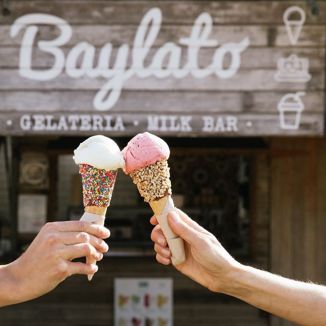 Two hands holding ice cream cones in front of a wooden sign that reads 'Bajlato' with smaller text below saying 'Gelateria, Milk Bar'. The cones have colorful sprinkles and chopped nuts.