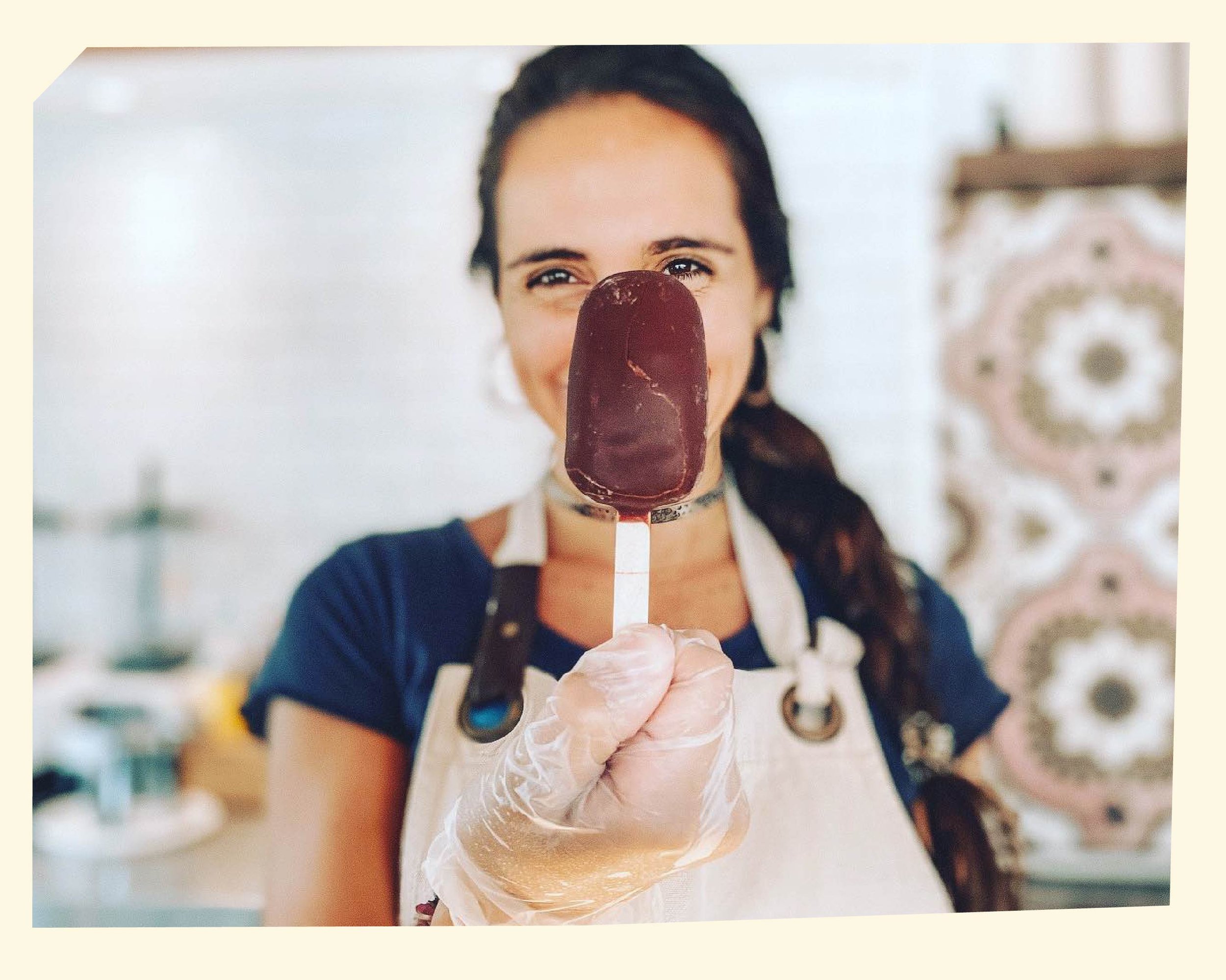 A woman holding a cherry popsicle and smiling at the camera in a kitchen, wearing a beige apron and plastic gloves.