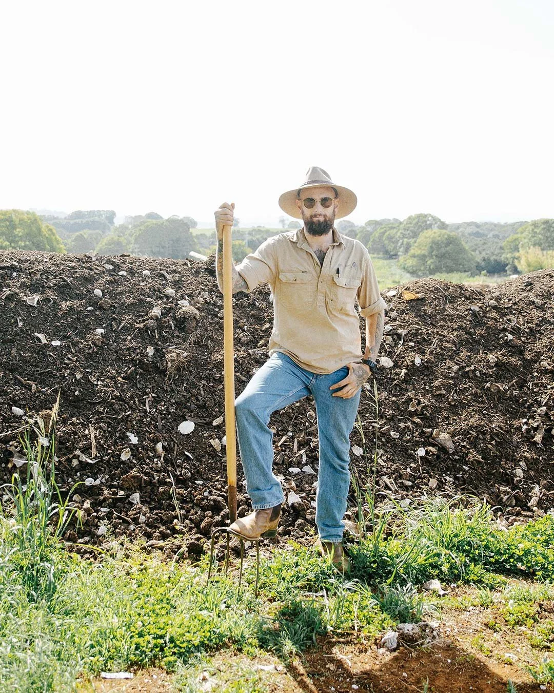 A man with a beard, wearing sunglasses, a wide-brimmed hat, a beige shirt, and jeans, standing outdoors near a mound of soil, holding a gardening fork in a natural landscape with trees and hills in the background.