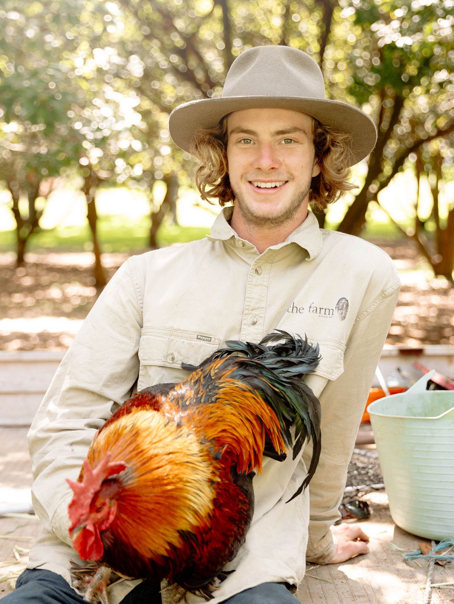 A smiling young man wearing a wide-brimmed hat and beige shirt holding a colorful rooster outdoors in a park or farm setting with trees and sunlight in the background.