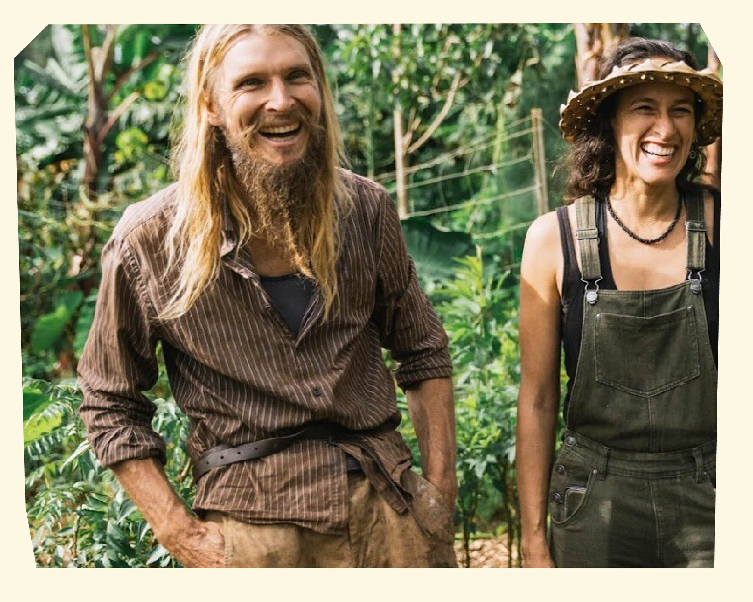 Smiling man with long blonde hair and beard, wearing a brown striped shirt, standing outdoors in a green jungle environment, next to a smiling woman with dark curly hair, wearing a wide-brimmed hat and overalls.