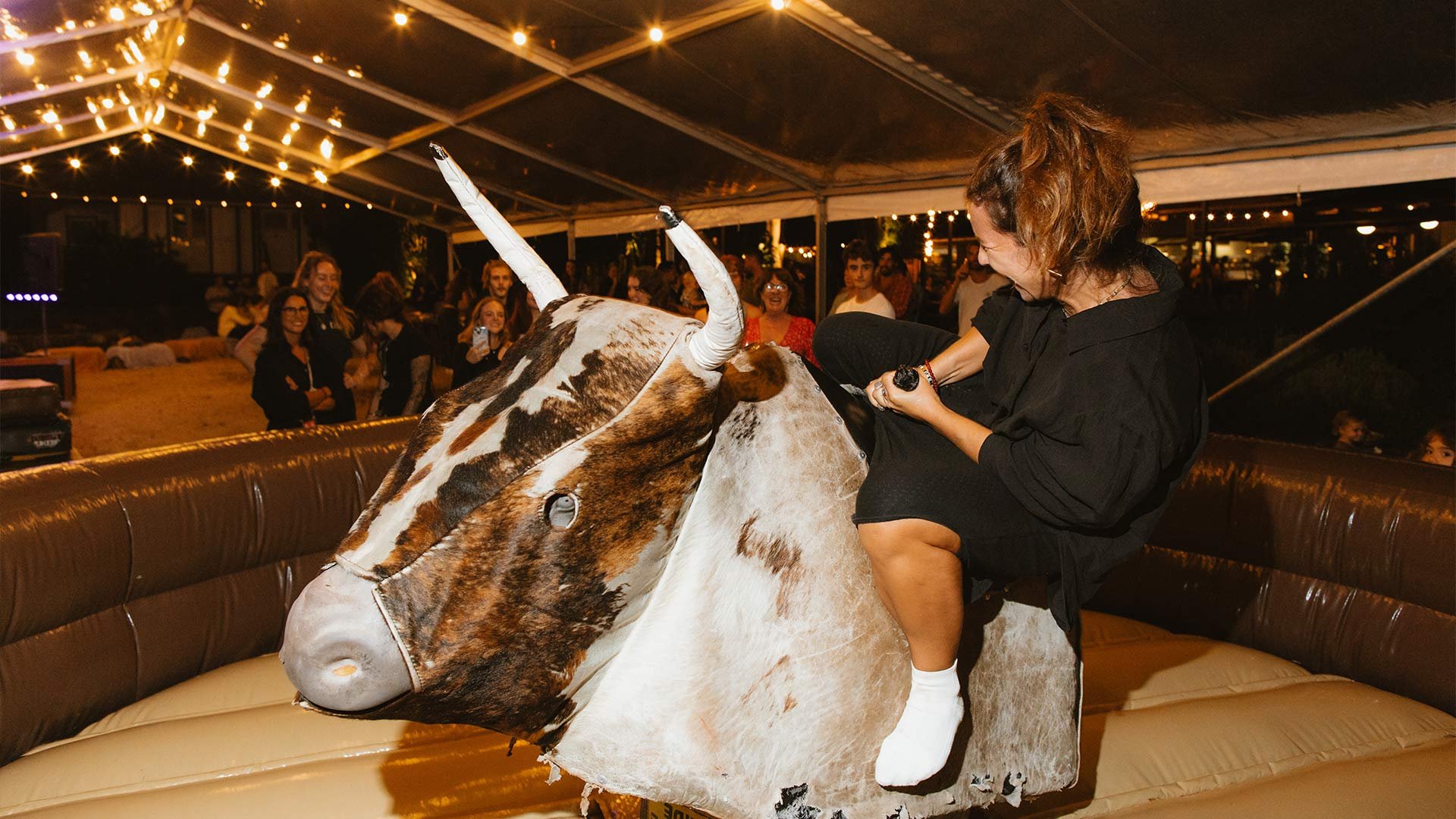 A woman riding a mechanical bull at a nighttime event with string lights overhead. She is smiling and holding the bull's horns while an audience watches and laughs in the background.