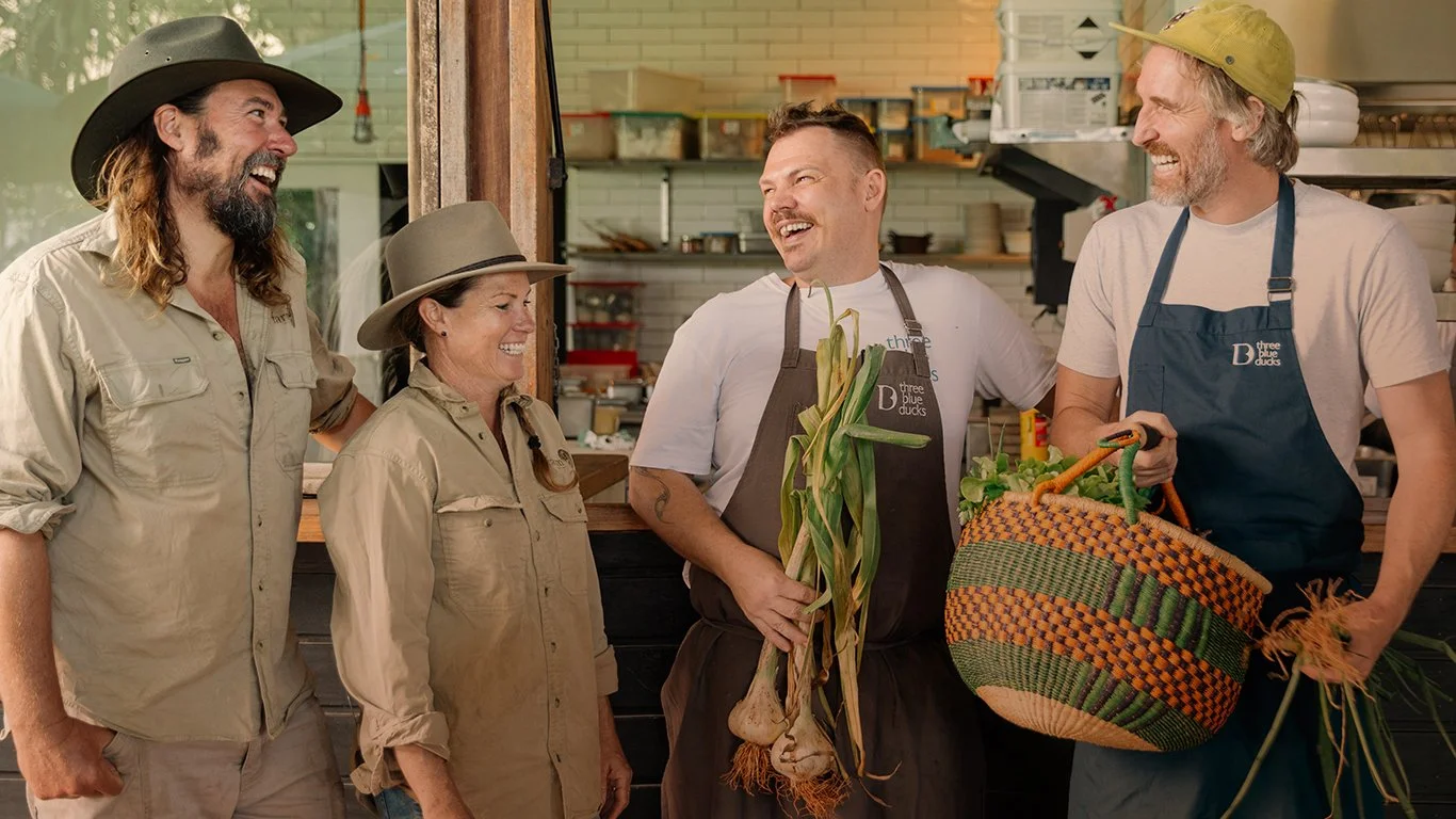 Four people laughing and talking inside a kitchen or market setting. They are wearing casual outdoor clothes and hats, and one person is holding a bunch of garlic and leek. Another person is holding a large basket filled with produce. All appear to be enjoying a friendly conversation.