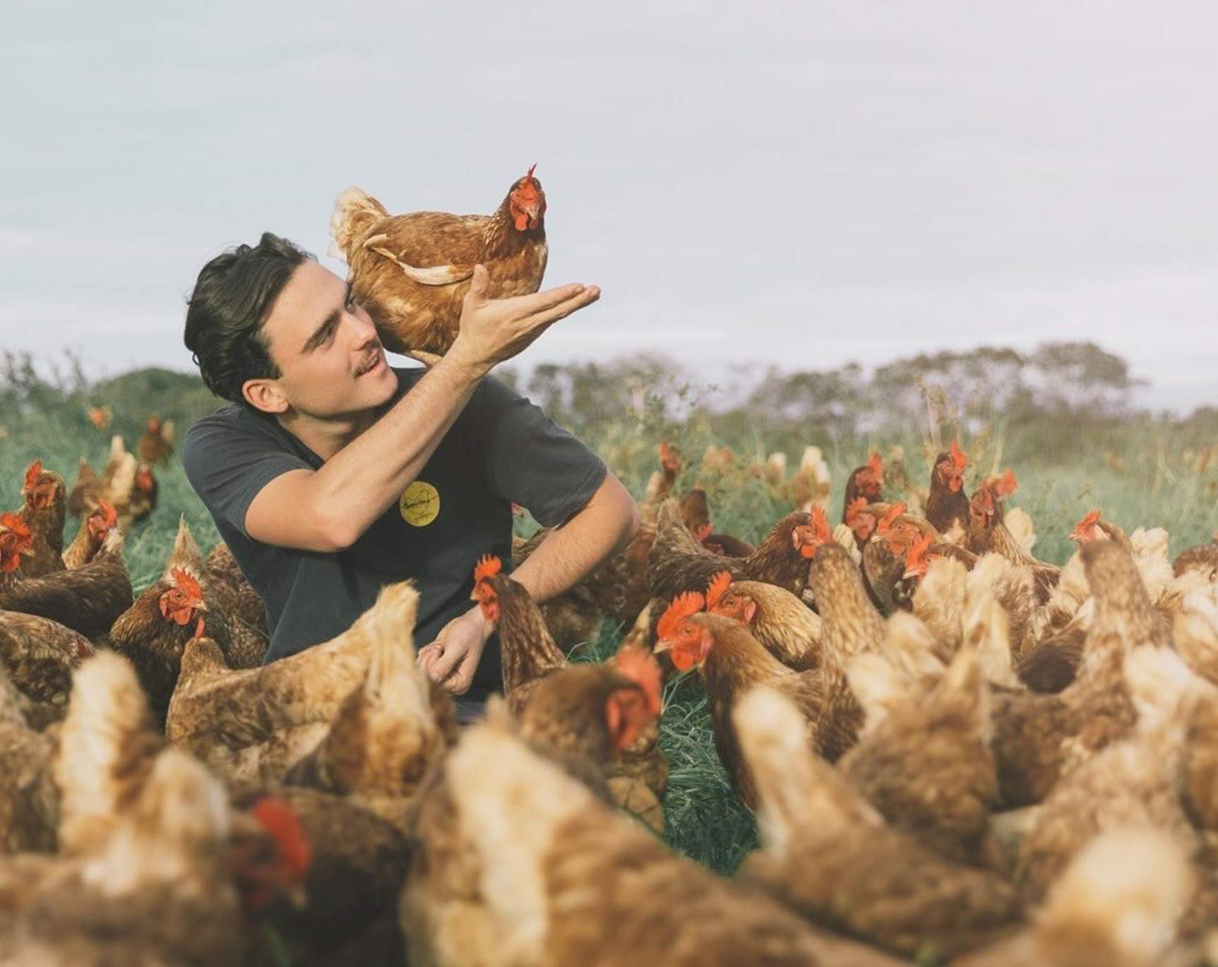 A young man sitting on the ground surrounded by chickens in a farm setting, holding a hen in his hand and smiling.