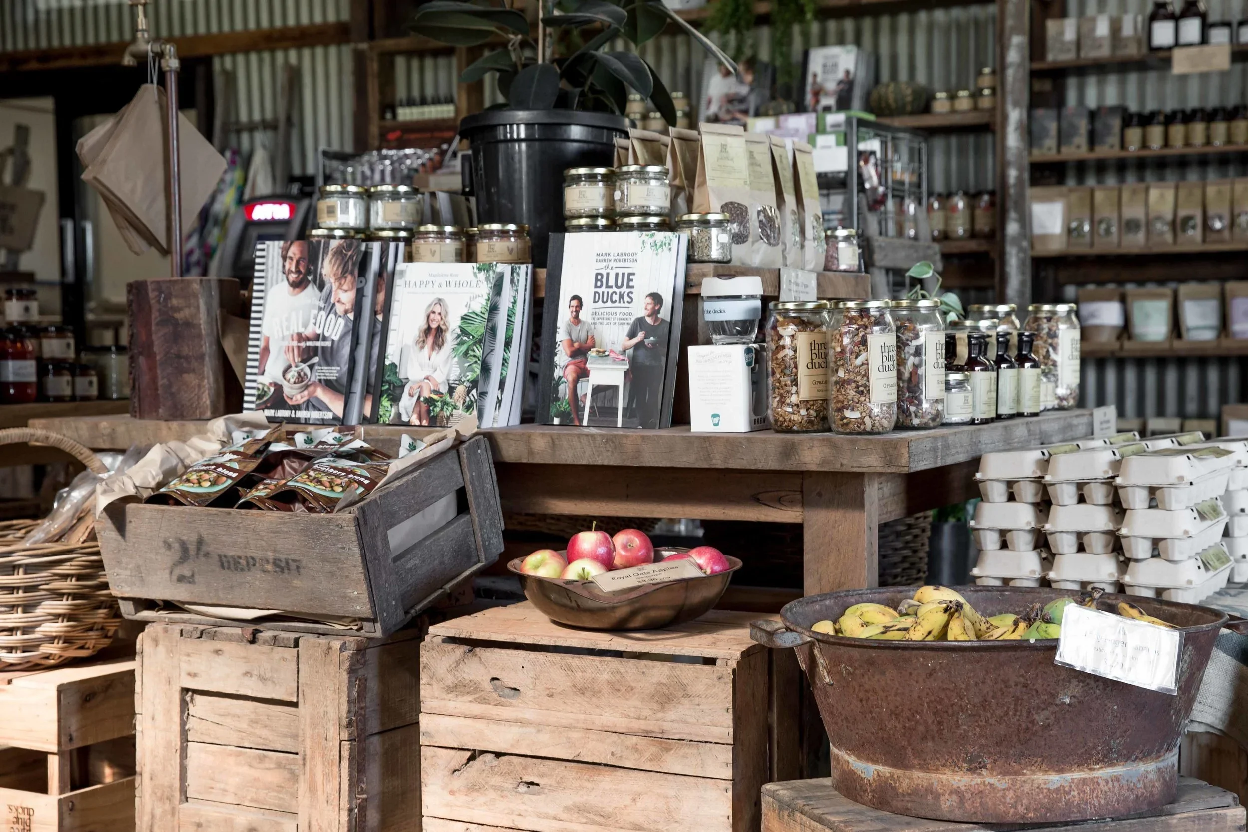 Display of jars, packaged food, and magazines on rustic wooden tables in a farm shop, with crates of apples and bananas below.
