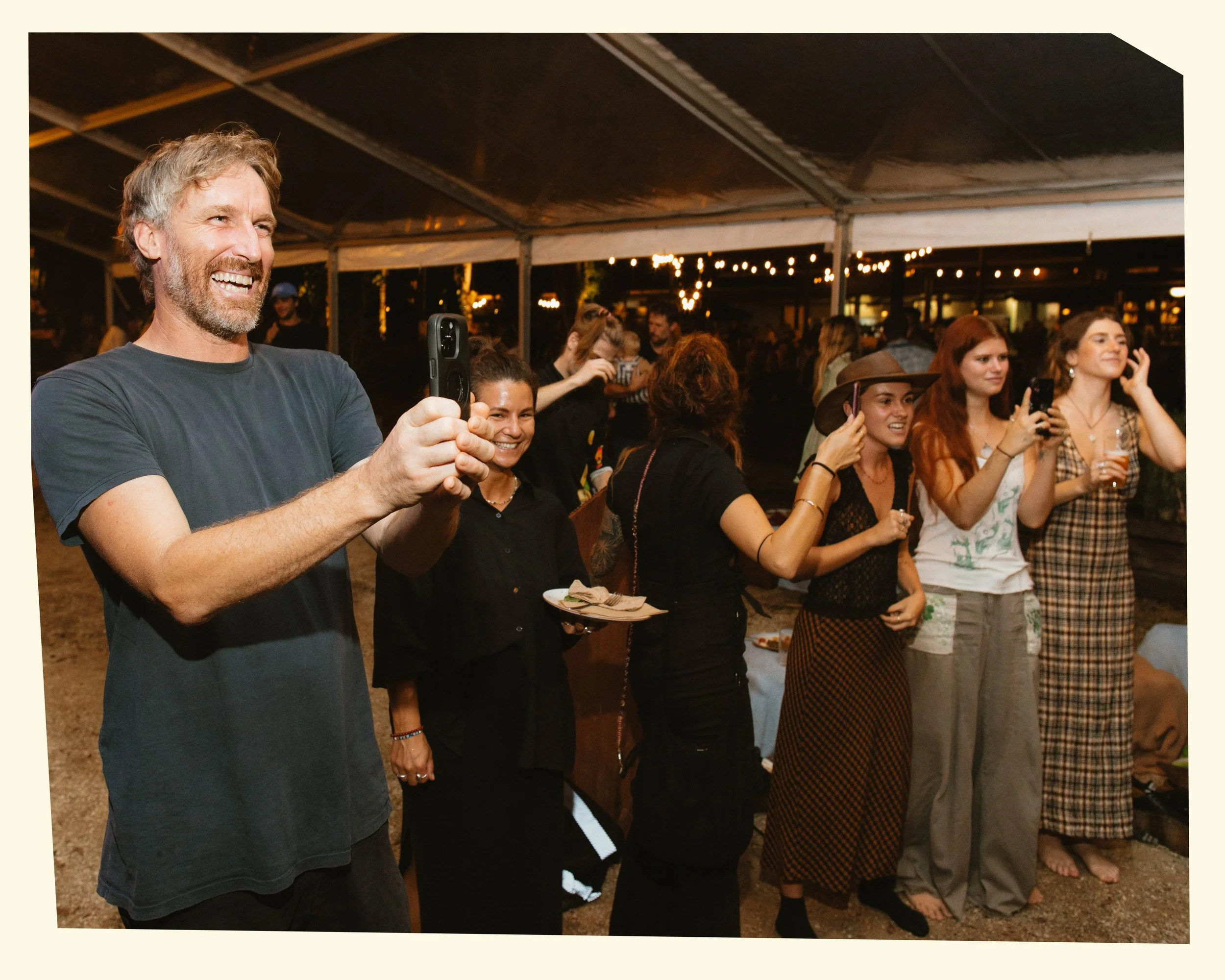 A group of people, mostly women, standing in line at an outdoor evening event, some holding drinks and using their phones, under string lights.