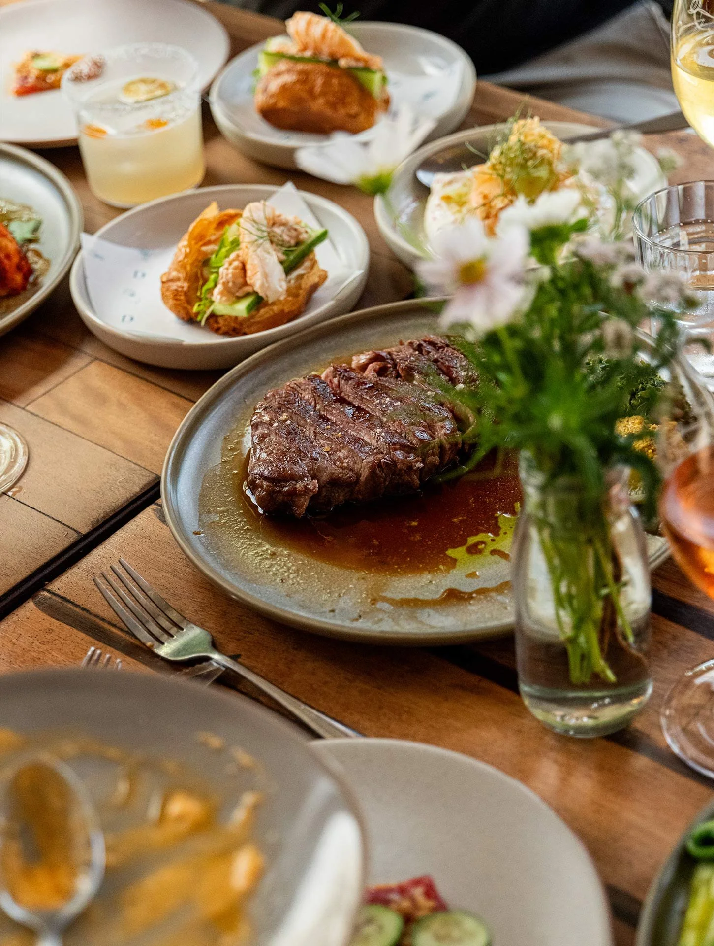 A table with various dishes including a large piece of cooked steak, small plates of appetizers, a glass of white wine, and a vase with pink flowers