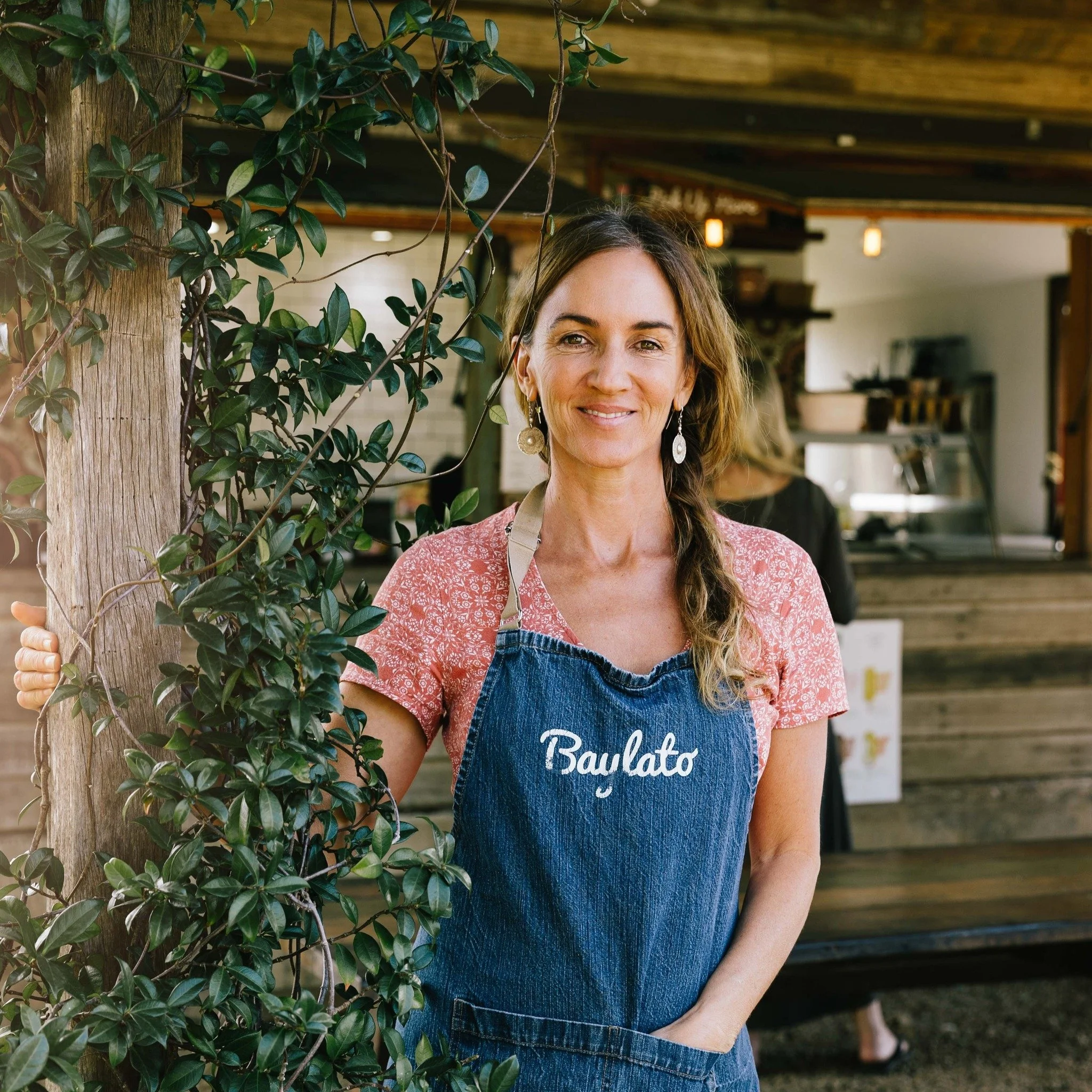 A woman with shoulder-length hair smiling, wearing earrings, a peach-colored top, blue apron, and standing outdoors behind a leafy tree.