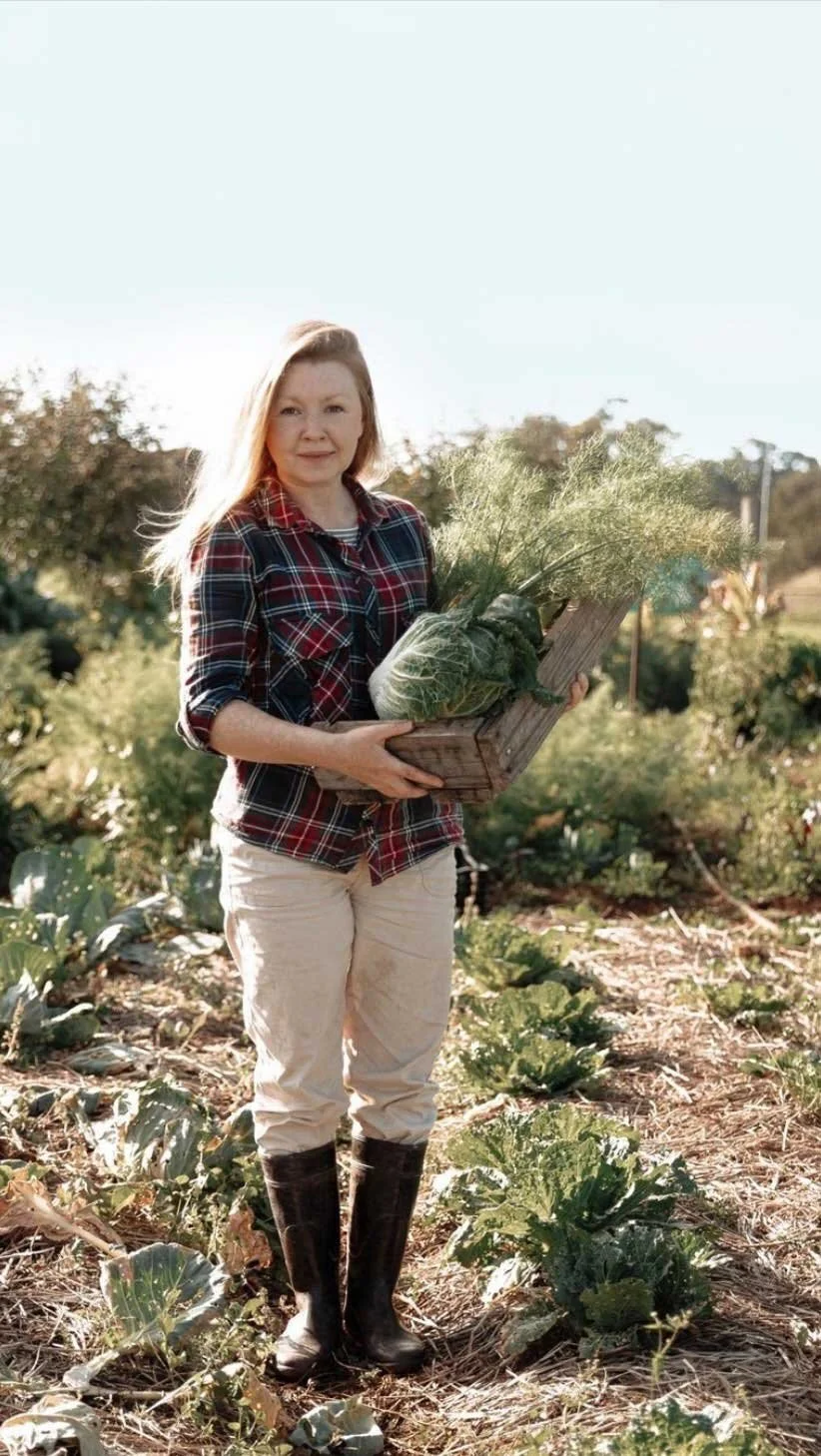 A woman standing in a farm field holding a wooden crate filled with fresh vegetables, including cabbage and leafy greens, wearing a plaid shirt, beige pants, and black boots.