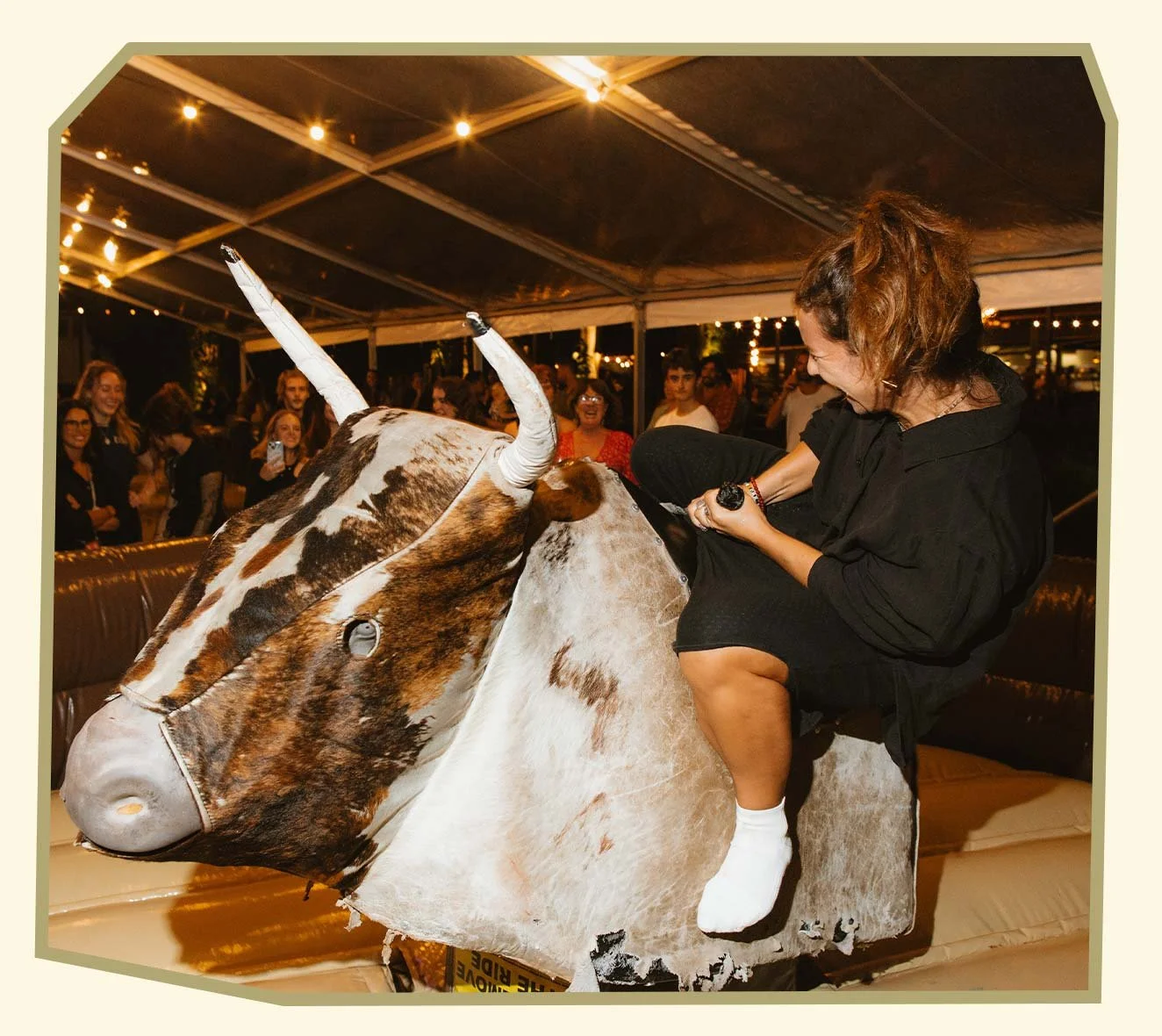 A woman riding a mechanical bull with a crowd of spectators watching and smiling in the background at night under string lights.