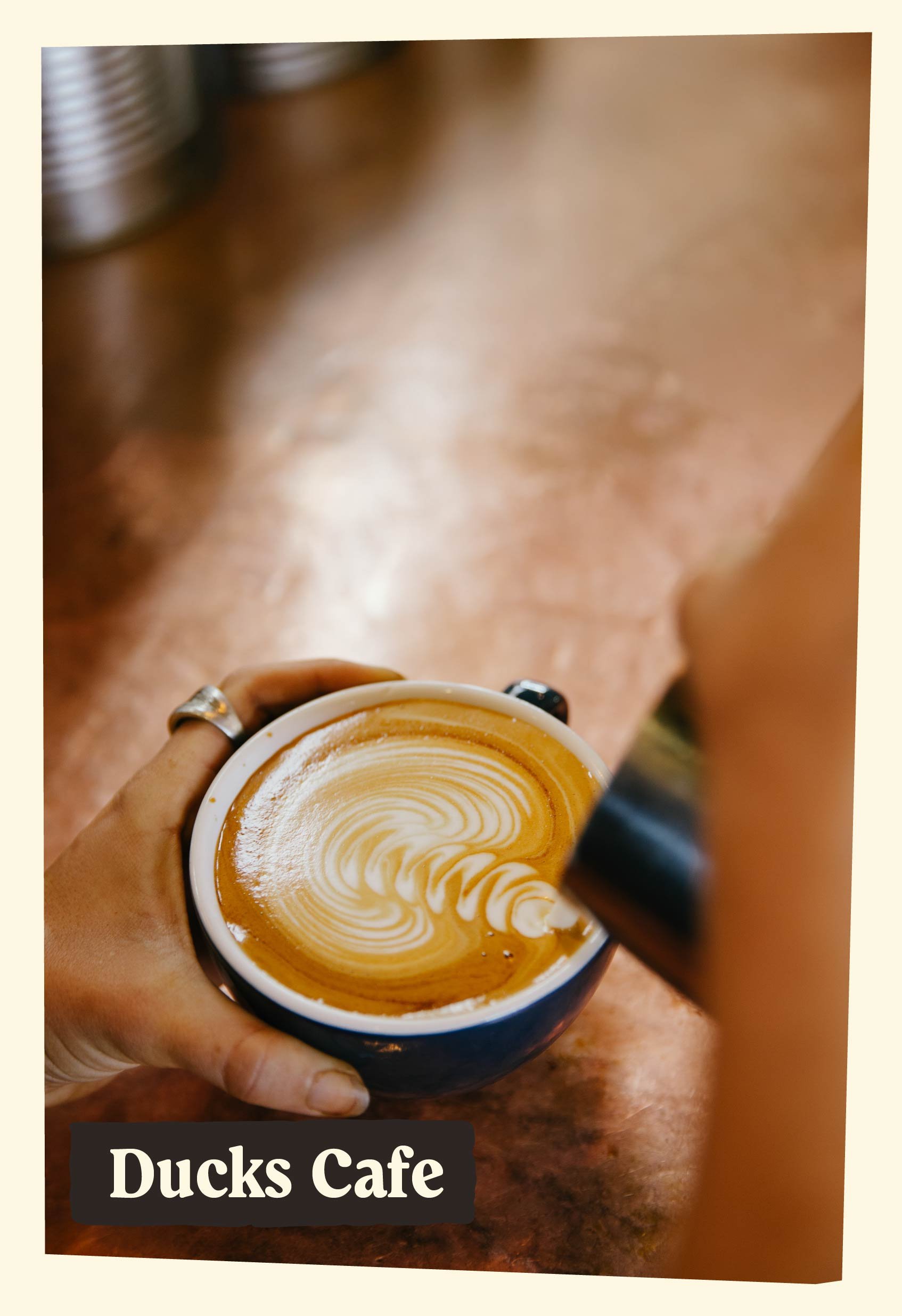 A person holding a cup of coffee with latte art, possibly from Ducks Cafe, with blurred background of a wooden table.