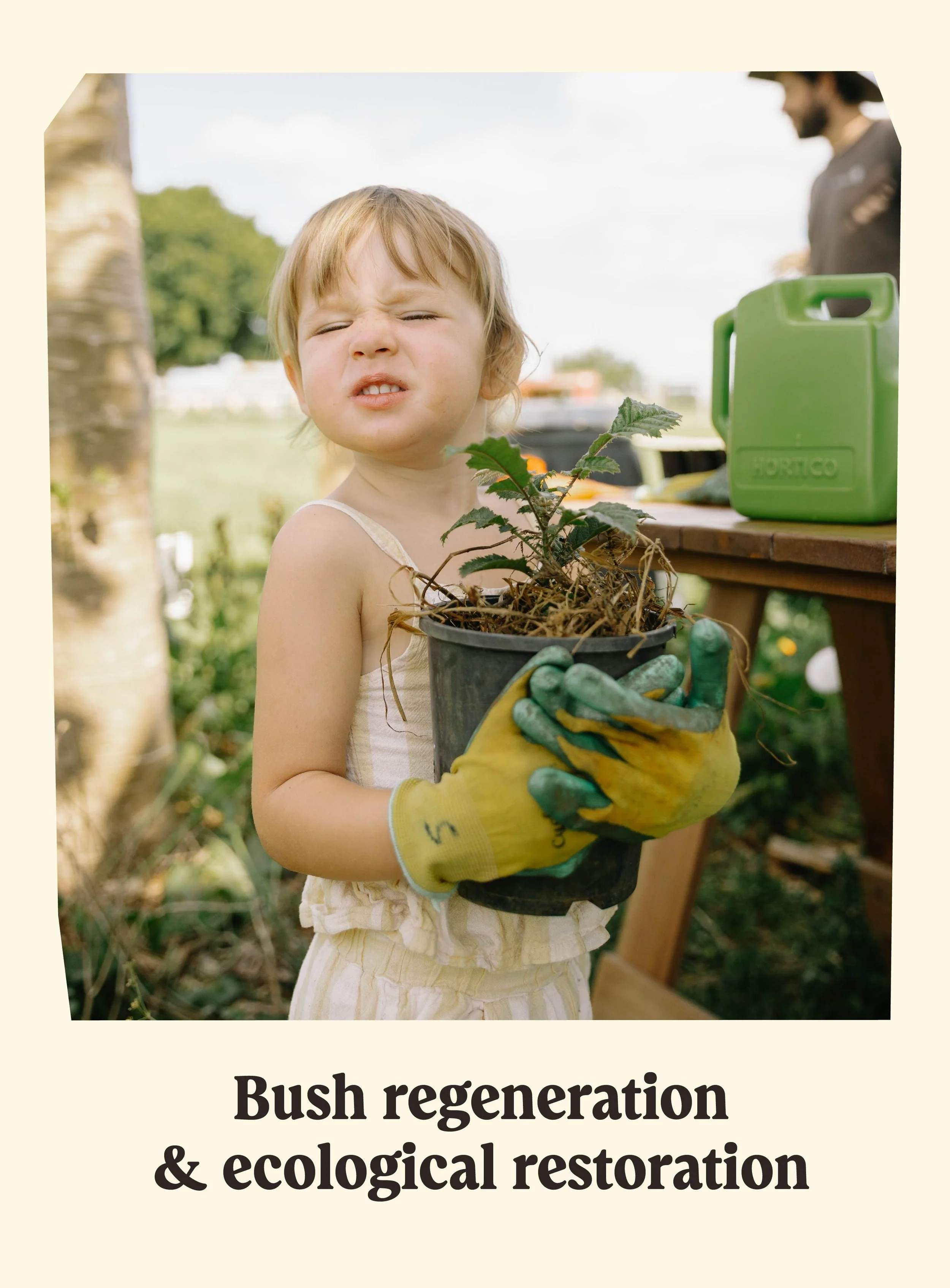 A young child with blond hair and a light-colored dress holds a pot of a small plant with green leaves, wearing yellow gardening gloves. In the background, a person in a hat is blurred, and there is outdoor scenery with a tree and a wooden table with gardening supplies. The scene appears to be an outdoor gardening or ecological restoration event.