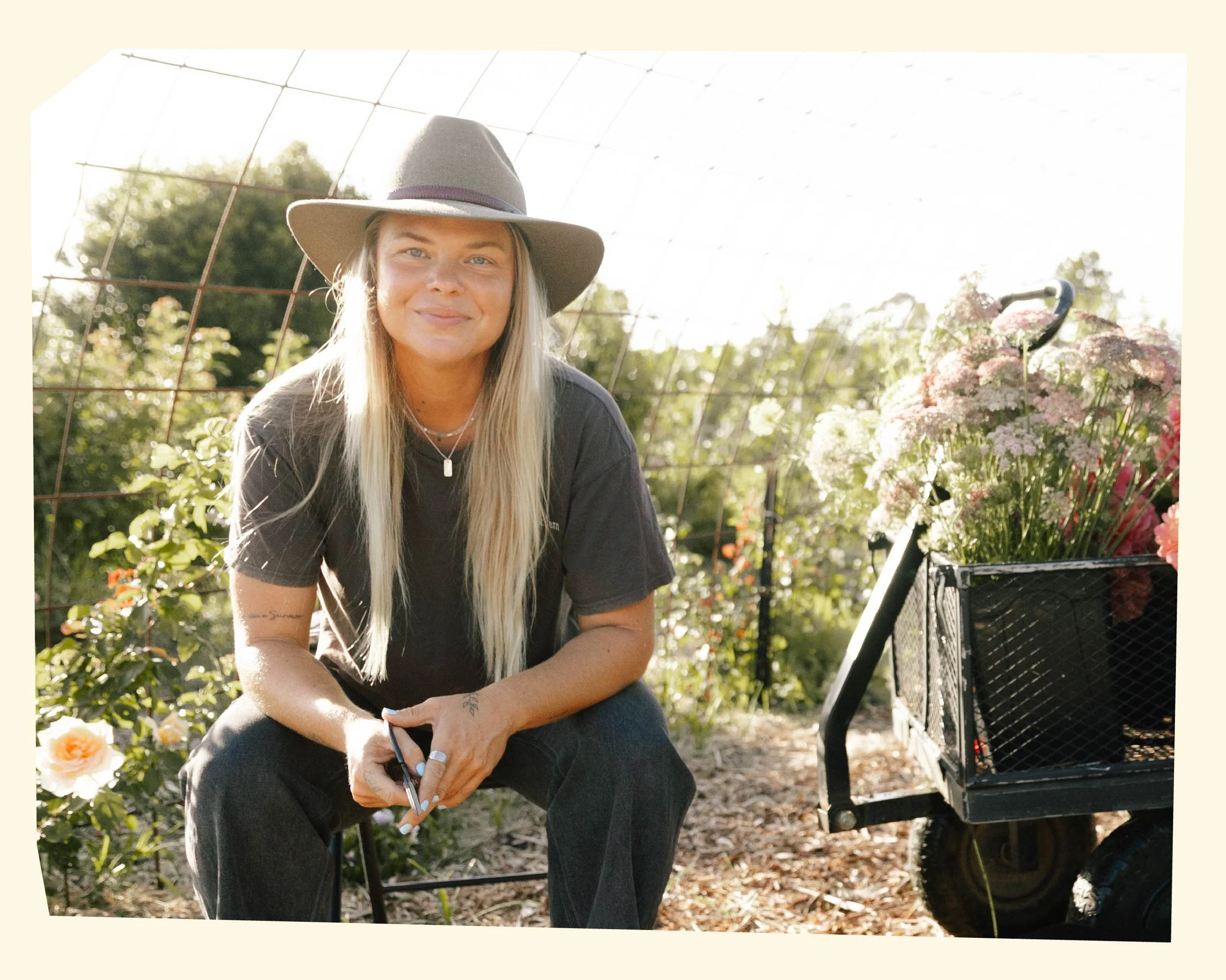 A woman with long blonde hair wearing a wide-brimmed hat and gray t-shirt sitting outdoors in a garden, with flowers and trees in the background and a cart filled with flowers beside her.